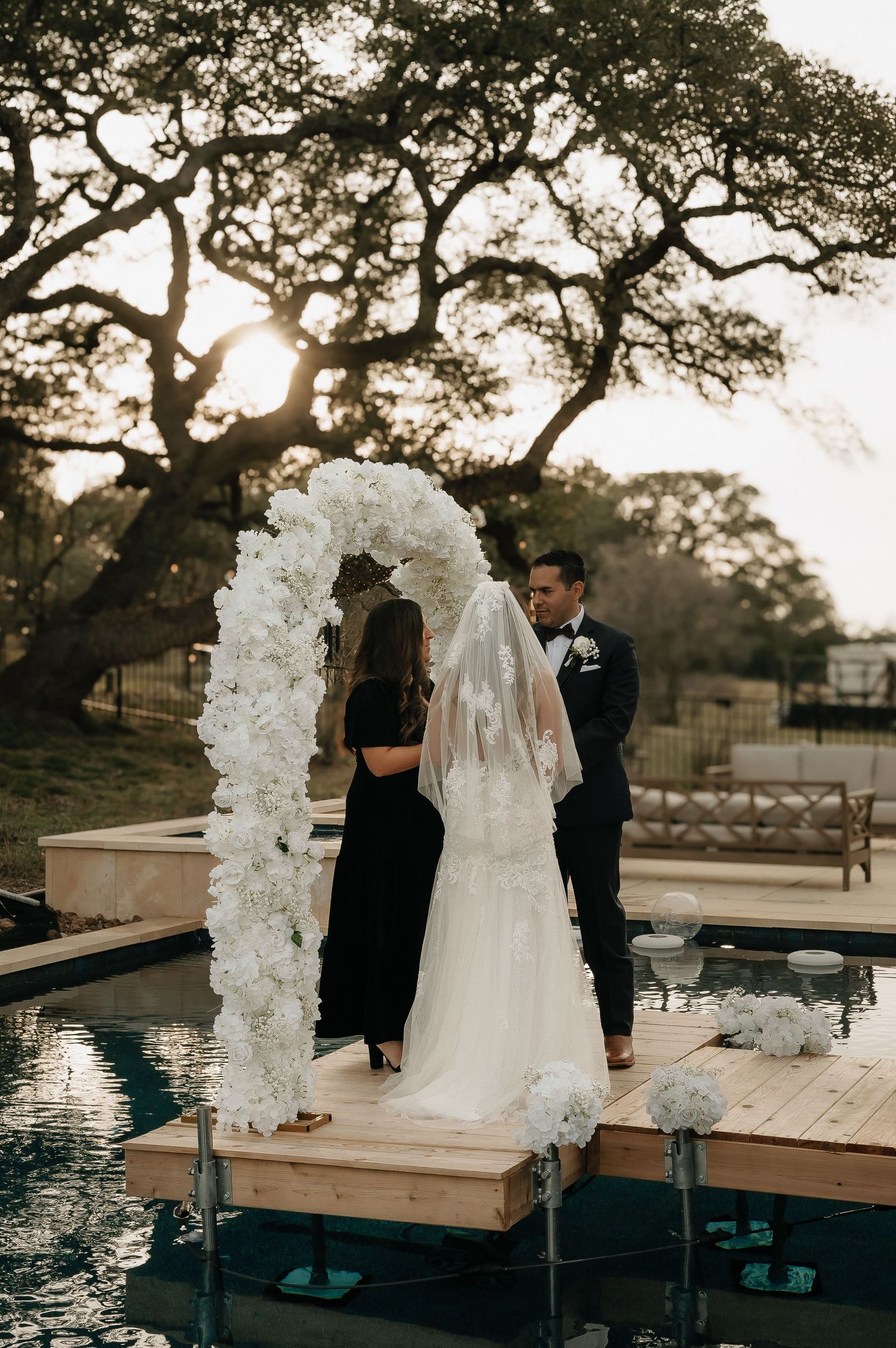 Outdoor wedding ceremony setup with white floral arch by a pool during golden hour.