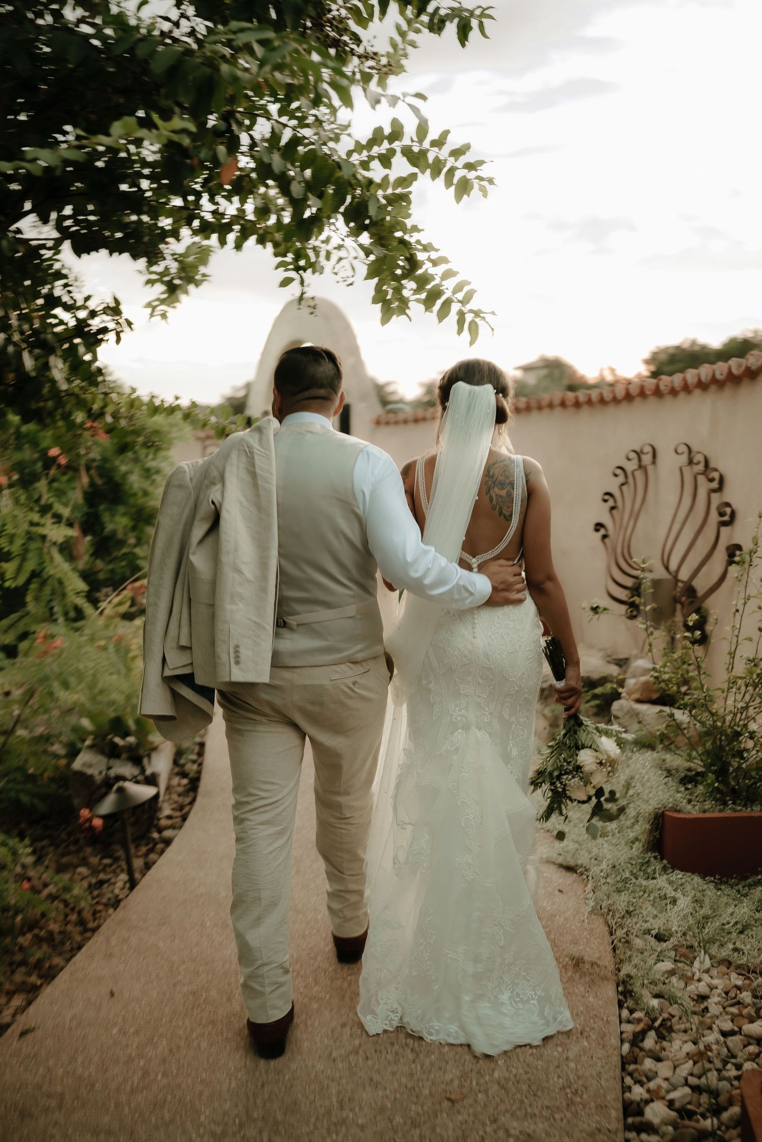 Newlyweds walking hand in hand along garden path at sunset.