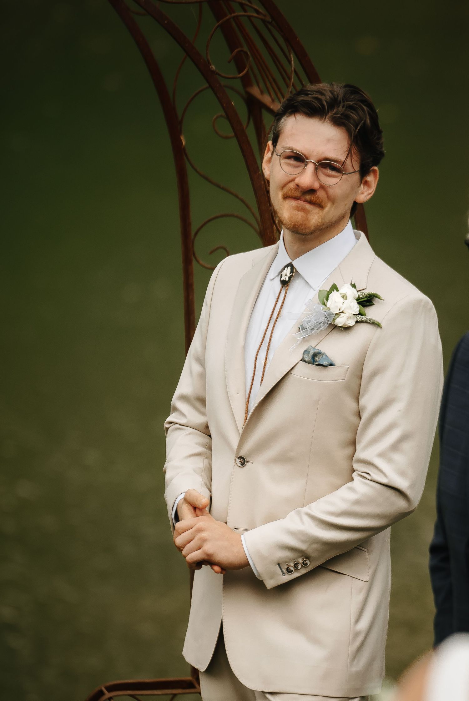 Groom in cream suit with bolo tie and boutonniere poses at outdoor wedding venue.