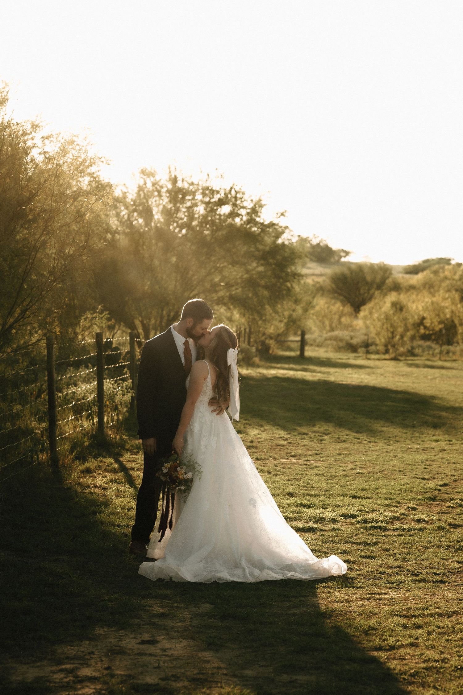 Newlyweds embrace in golden sunset light on grassy hillside.