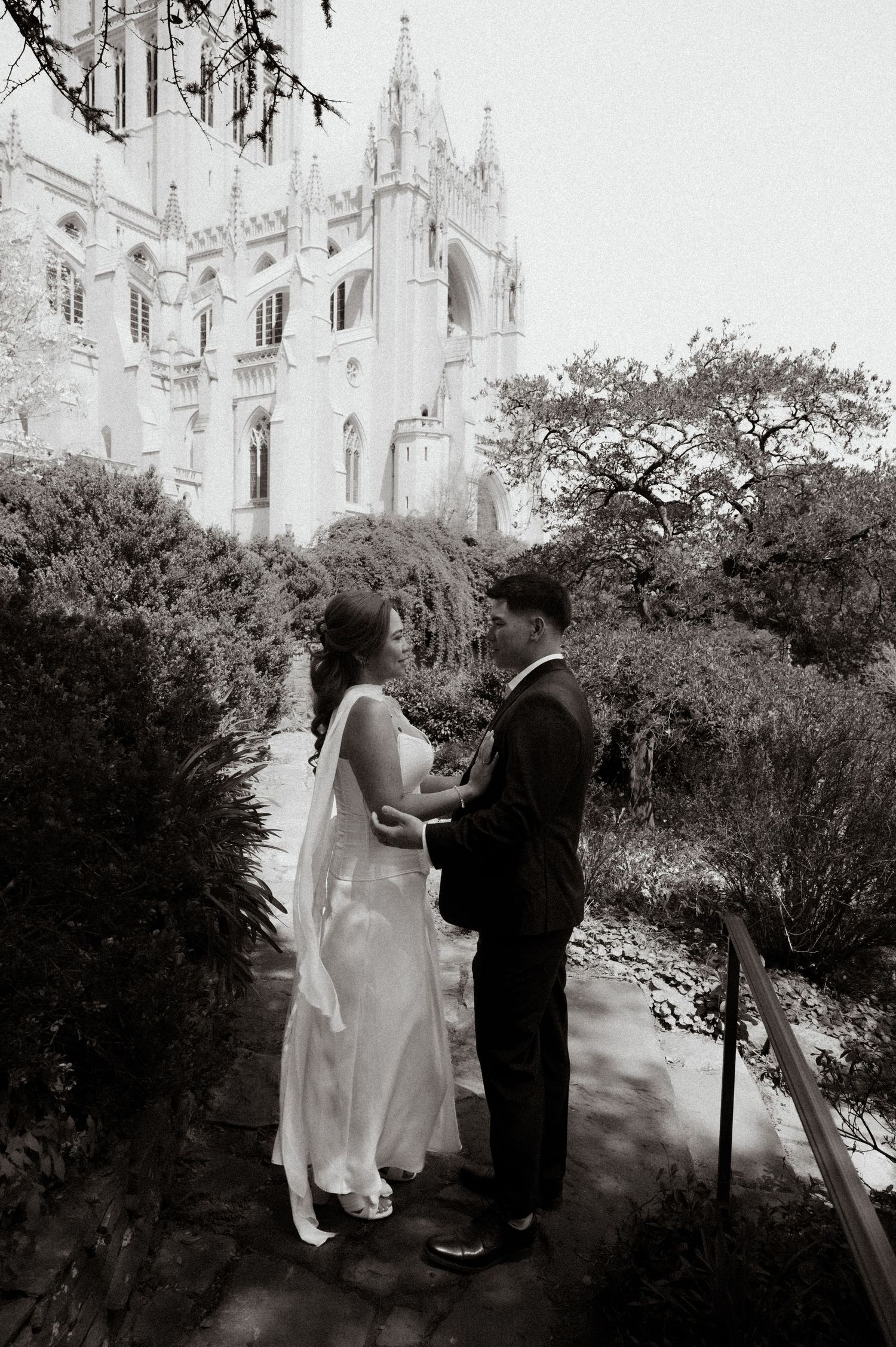 Black and white photo of a couple in wedding attire holding hands in a garden with a Gothic cathedral in the background.