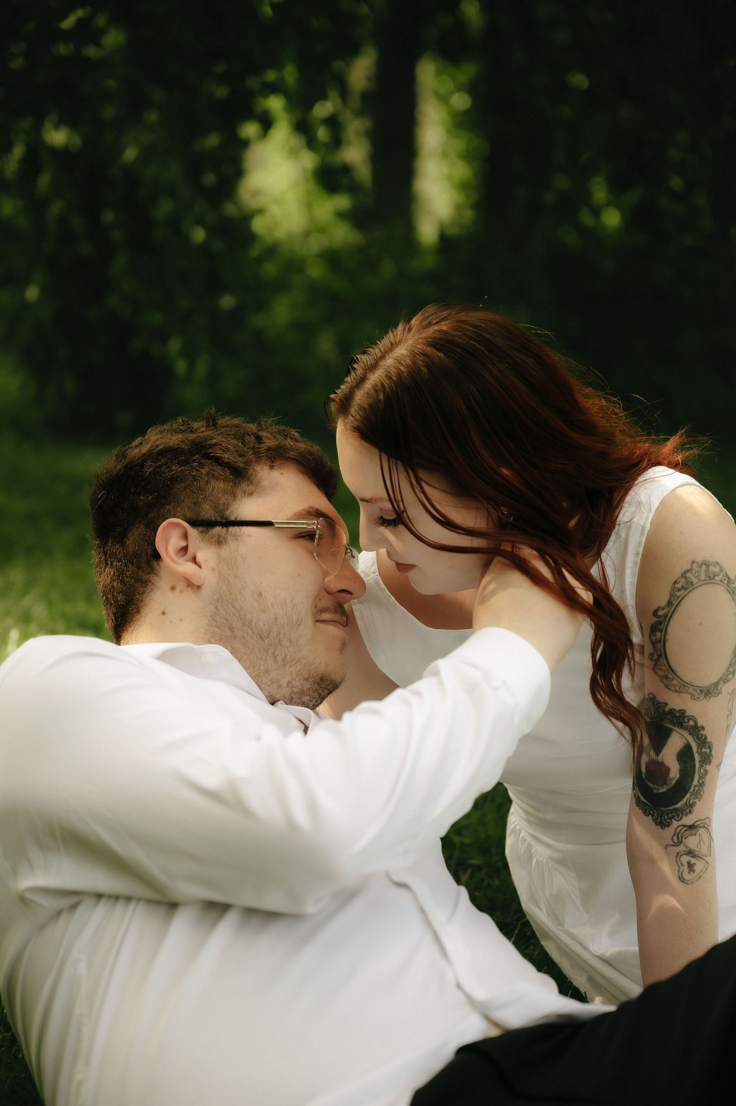 A couple shares a romantic moment outdoors, with a tattooed redhead leaning close to a man in glasses and white shirt.