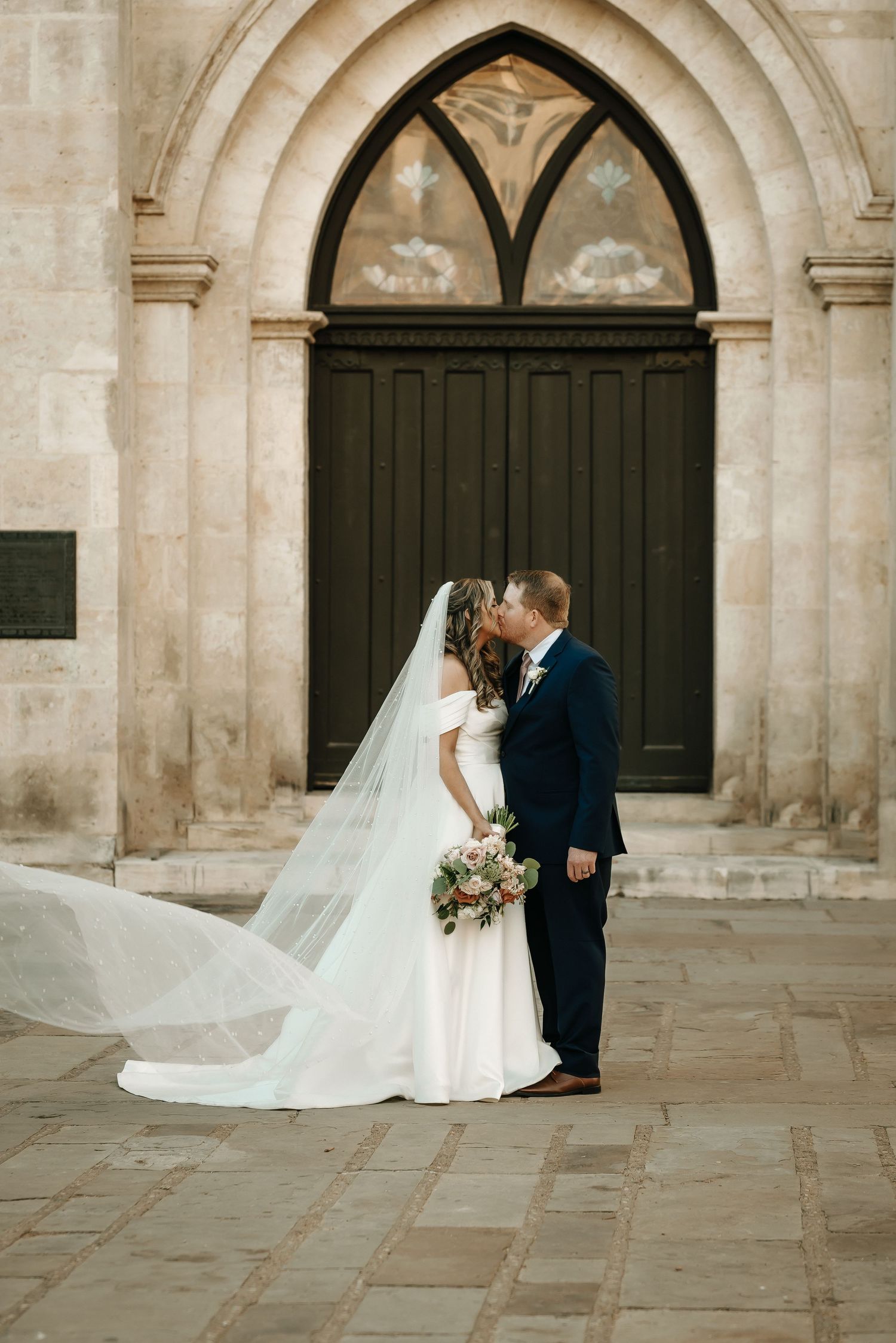 Newlyweds share a kiss in front of gothic stone archways.