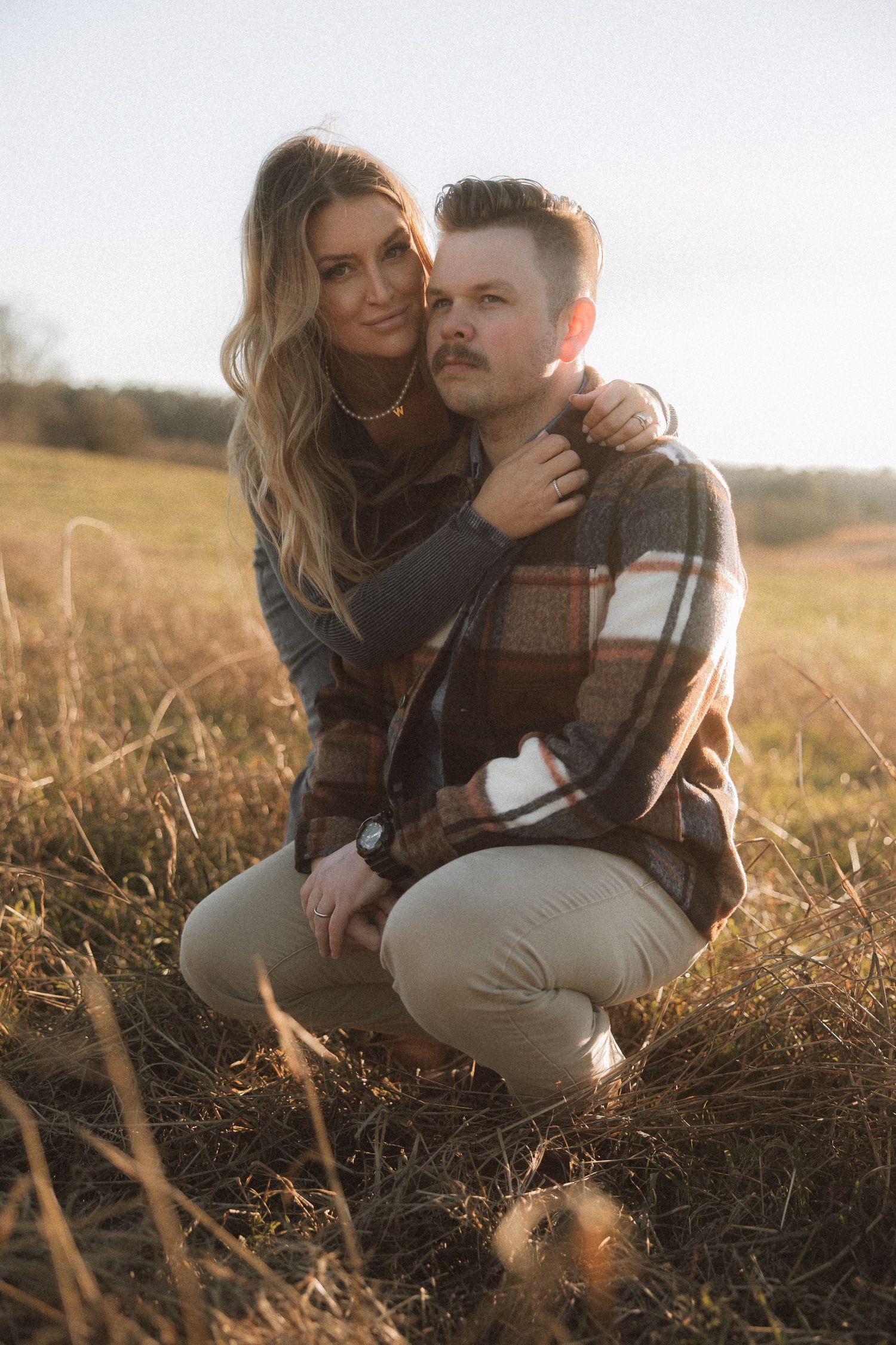A romantic couple sits together in a sunlit field during golden hour.