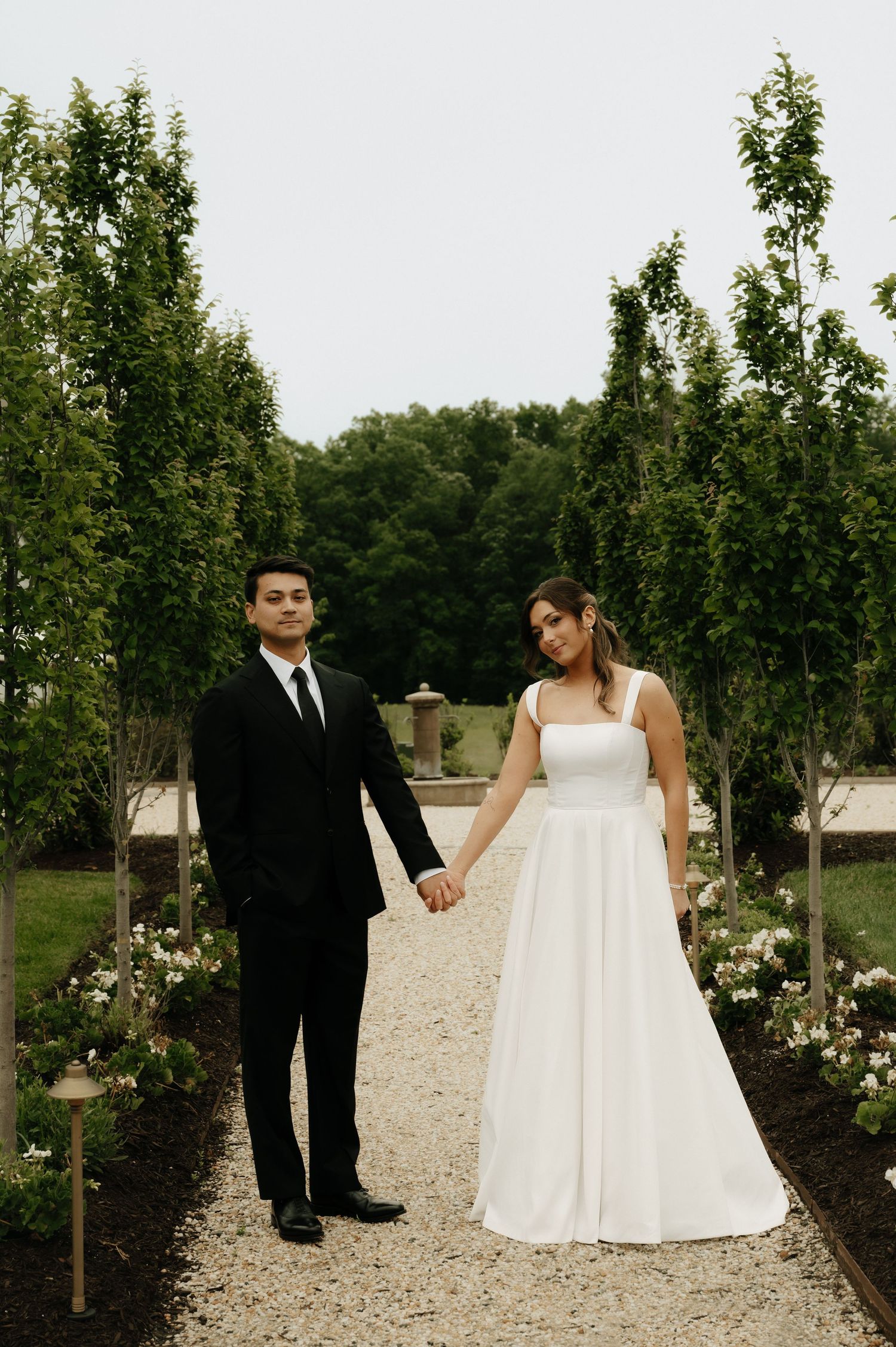 A bride in a white gown and groom in a black suit hold hands on a garden path lined with trees.