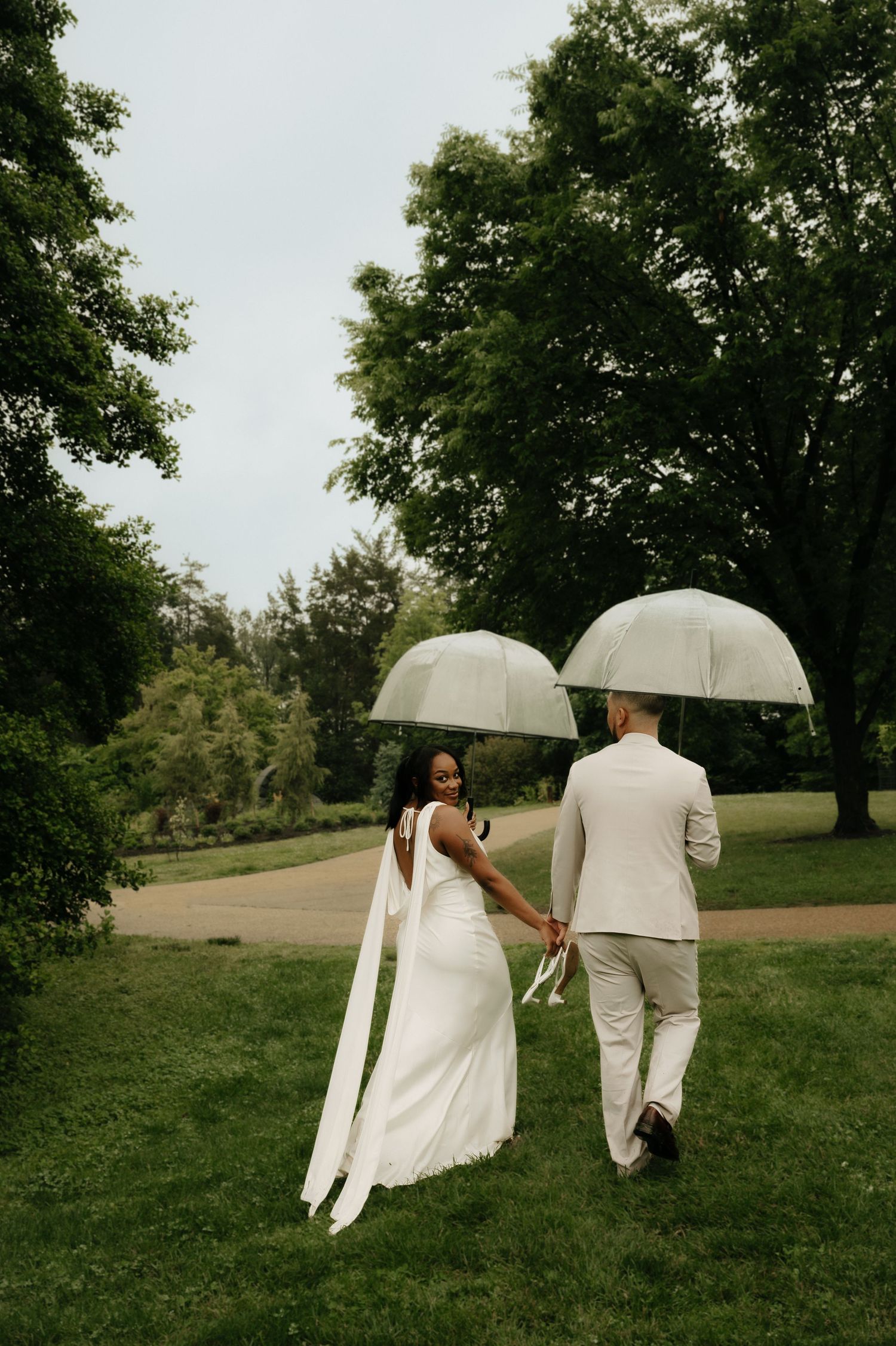 Bride and groom walking hand in hand through lush green park holding clear dome umbrellas on their wedding day.