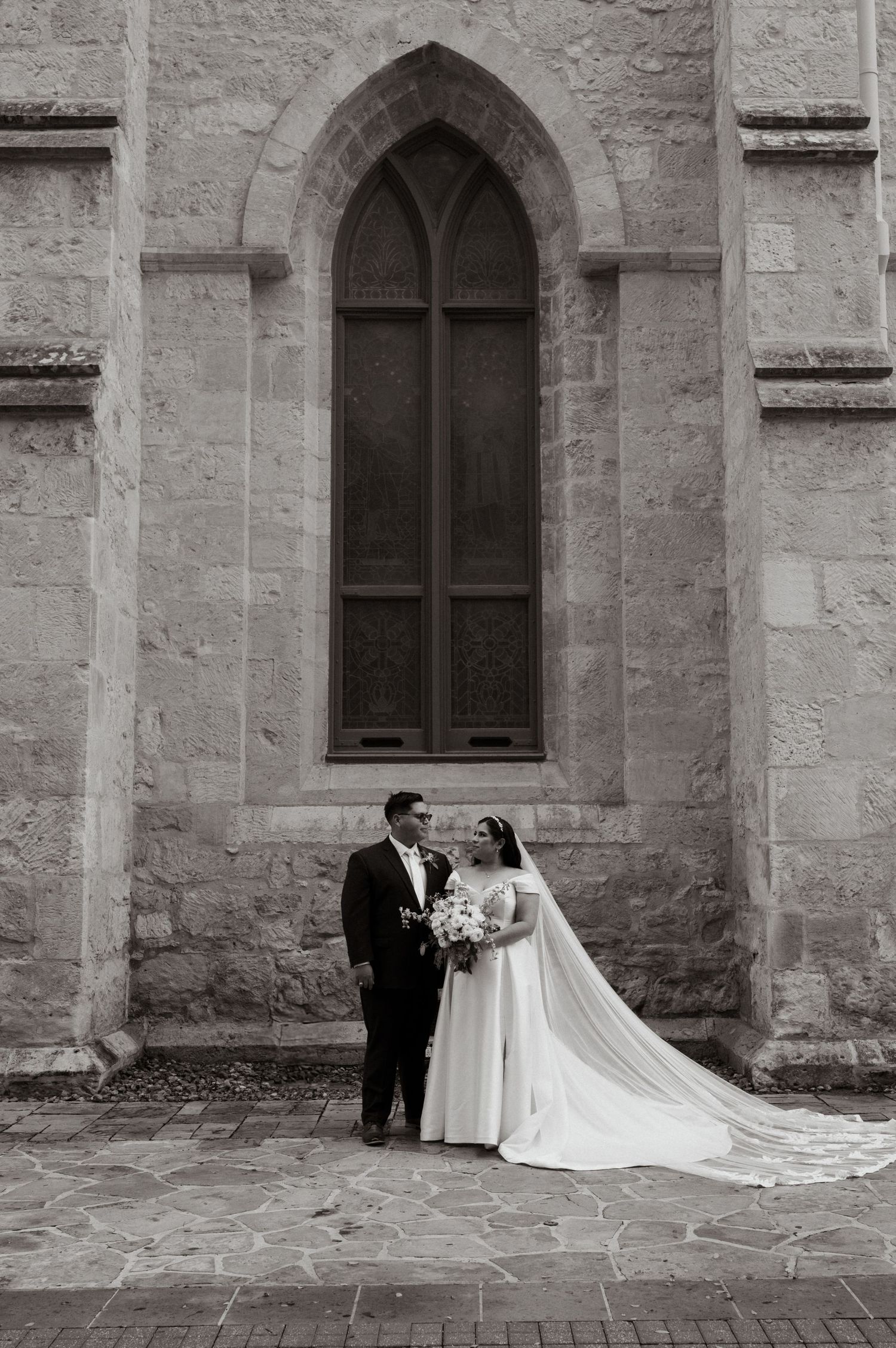 Black and white wedding photos against a stone church wall with elegant arched windows and flowing white dress train.