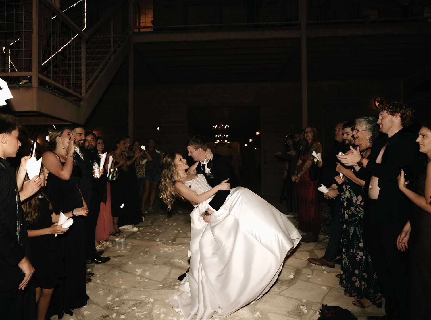 Wedding guests dance and celebrate on a dimly lit dance floor at an evening reception.
