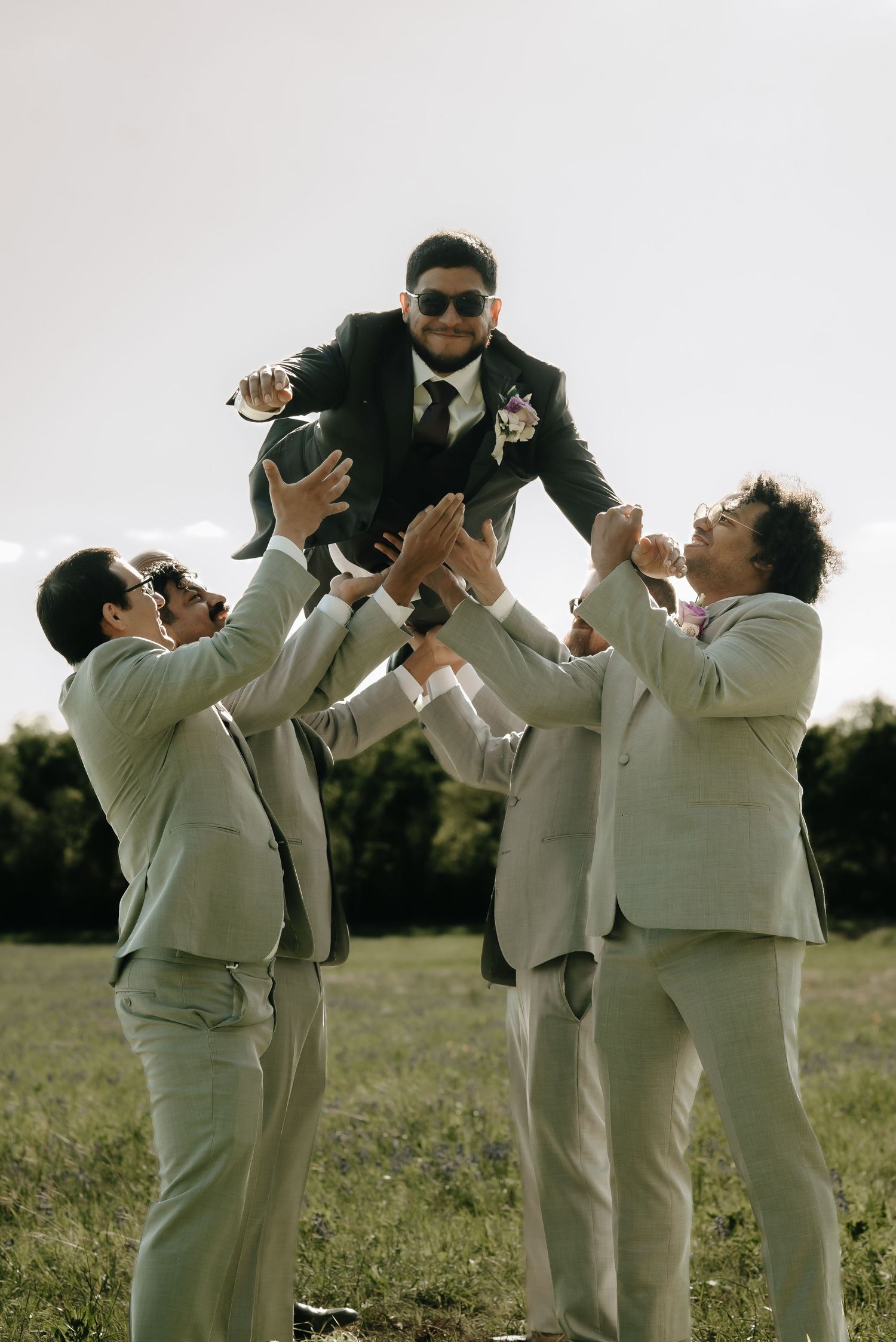 Groomsmen in light suits taking celebratory drinks in an outdoor field.