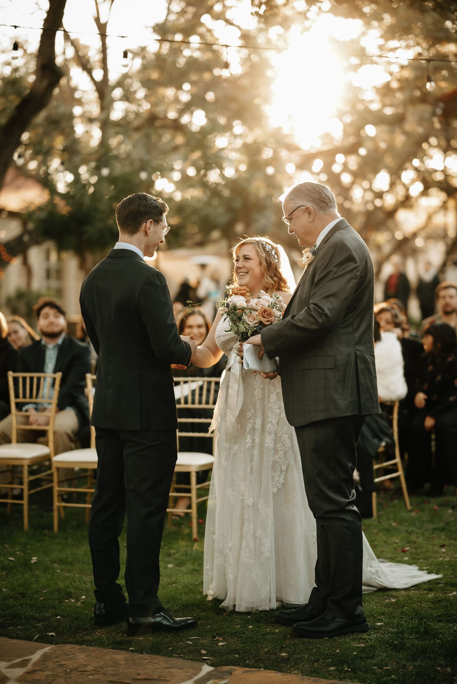 Outdoor wedding ceremony under golden sunlight with guests seated on gold chairs.