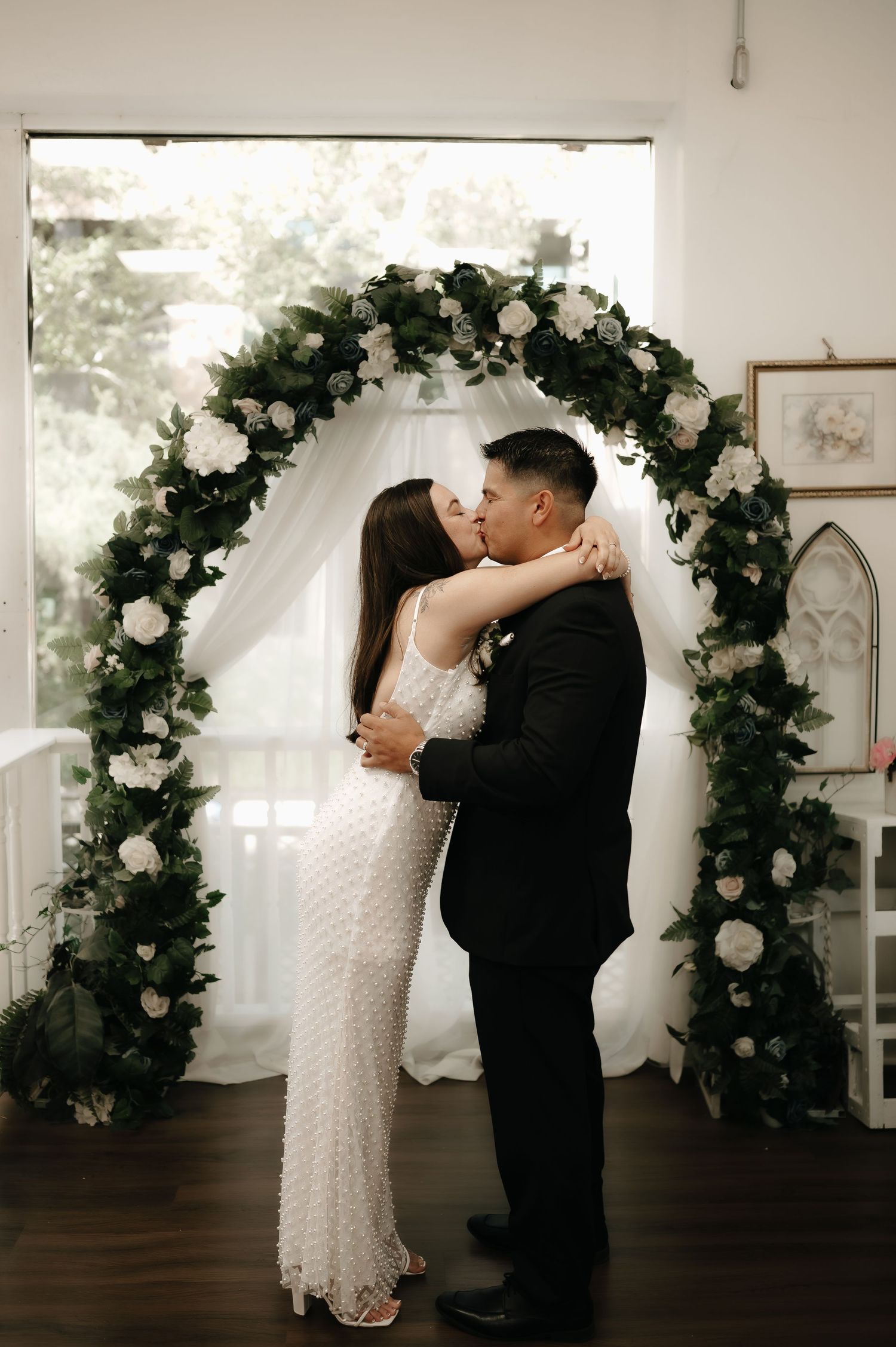 Wedding ceremony sequence under a floral arch with white roses and greenery in an indoor venue.