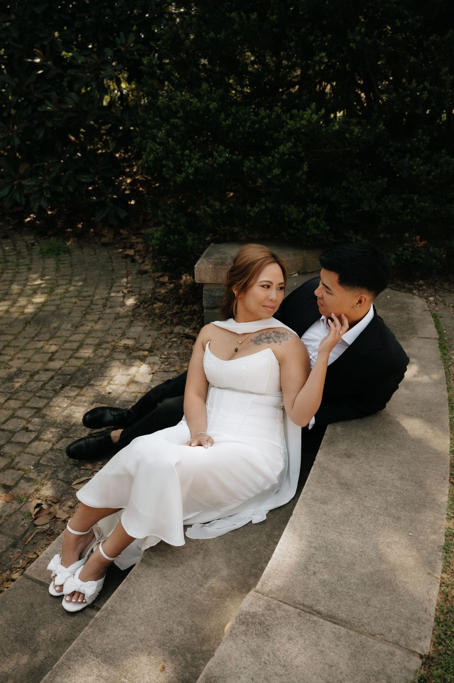 A bride in a white dress and groom in a black suit sit together on stone steps, gazing at each other in a garden.
