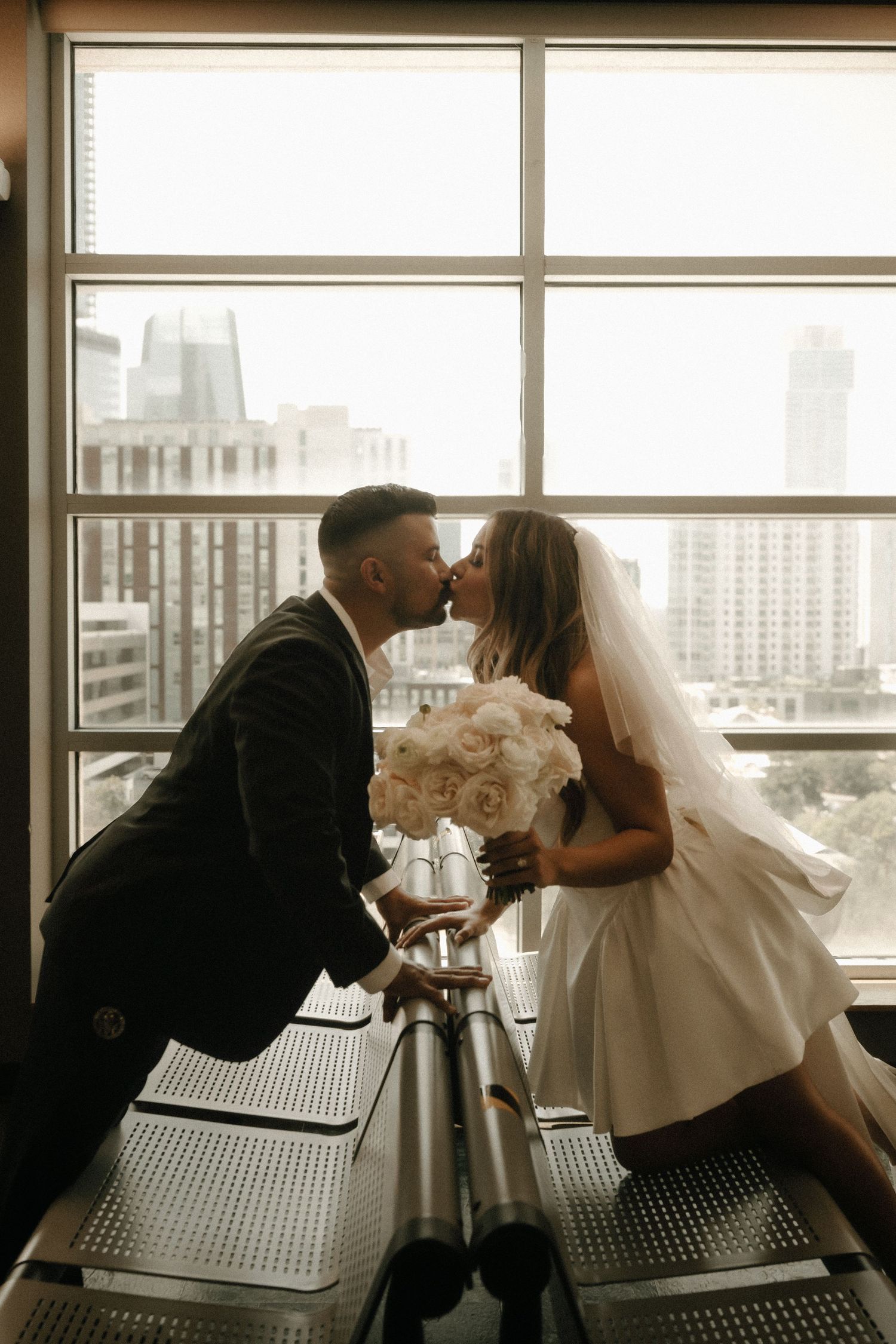 A romantic silhouette of newlyweds kissing by a large window with a cityscape view.