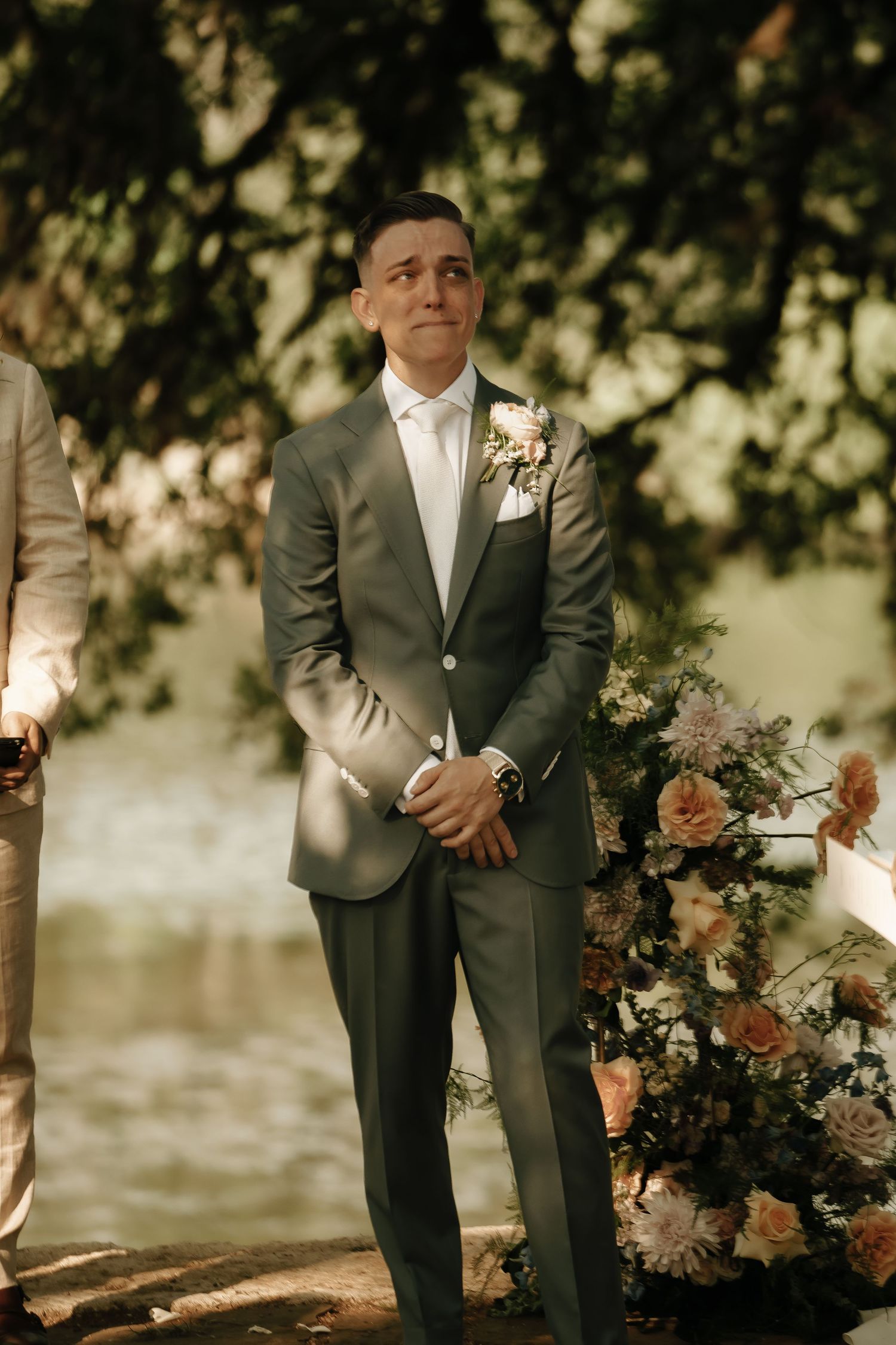 A groom in a gray suit stands in soft natural lighting under trees during an outdoor wedding ceremony.