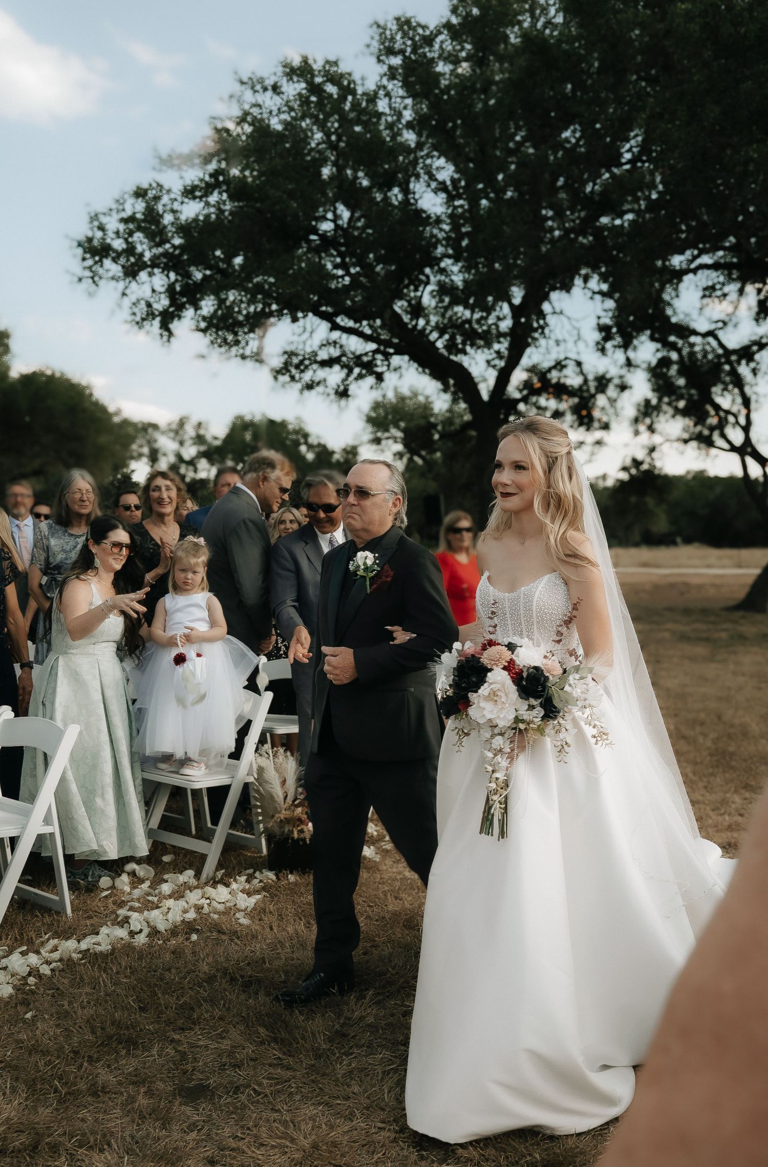 Wedding ceremony guests gather outdoors under oak trees with white chairs and floral decor.