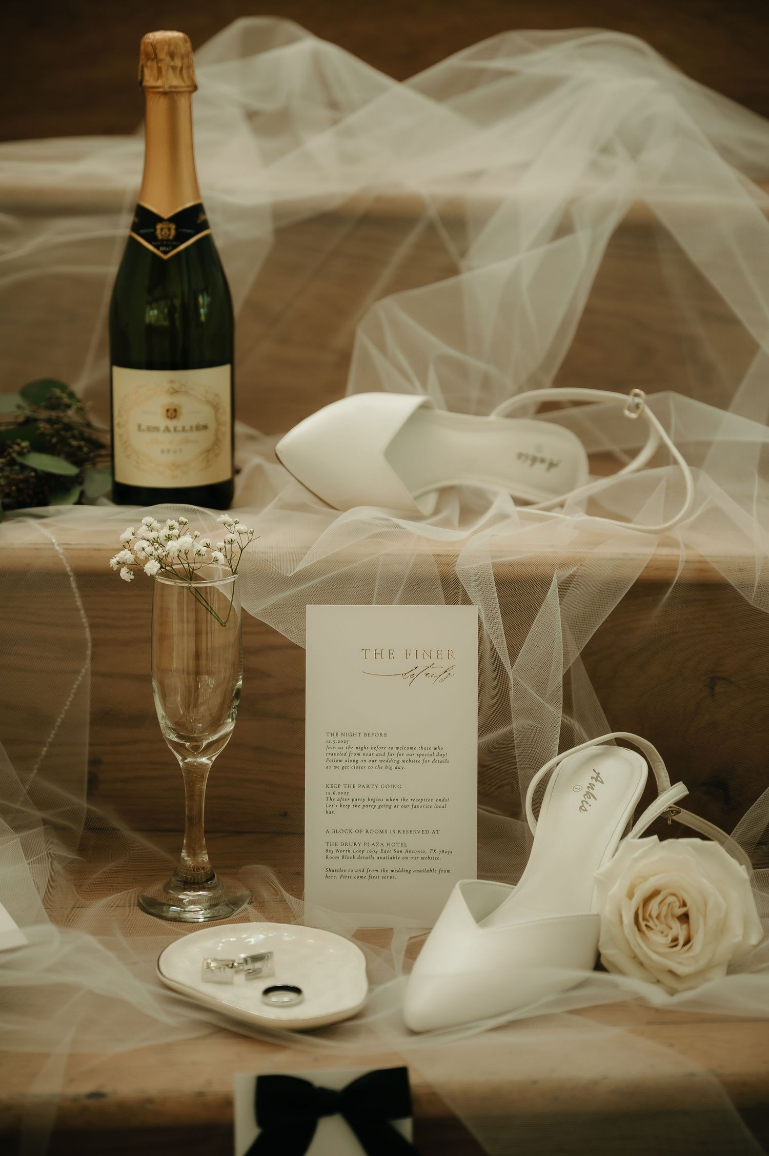 Wedding details including champagne bottles, tulle veil, and delicate roses arranged on wooden surface.