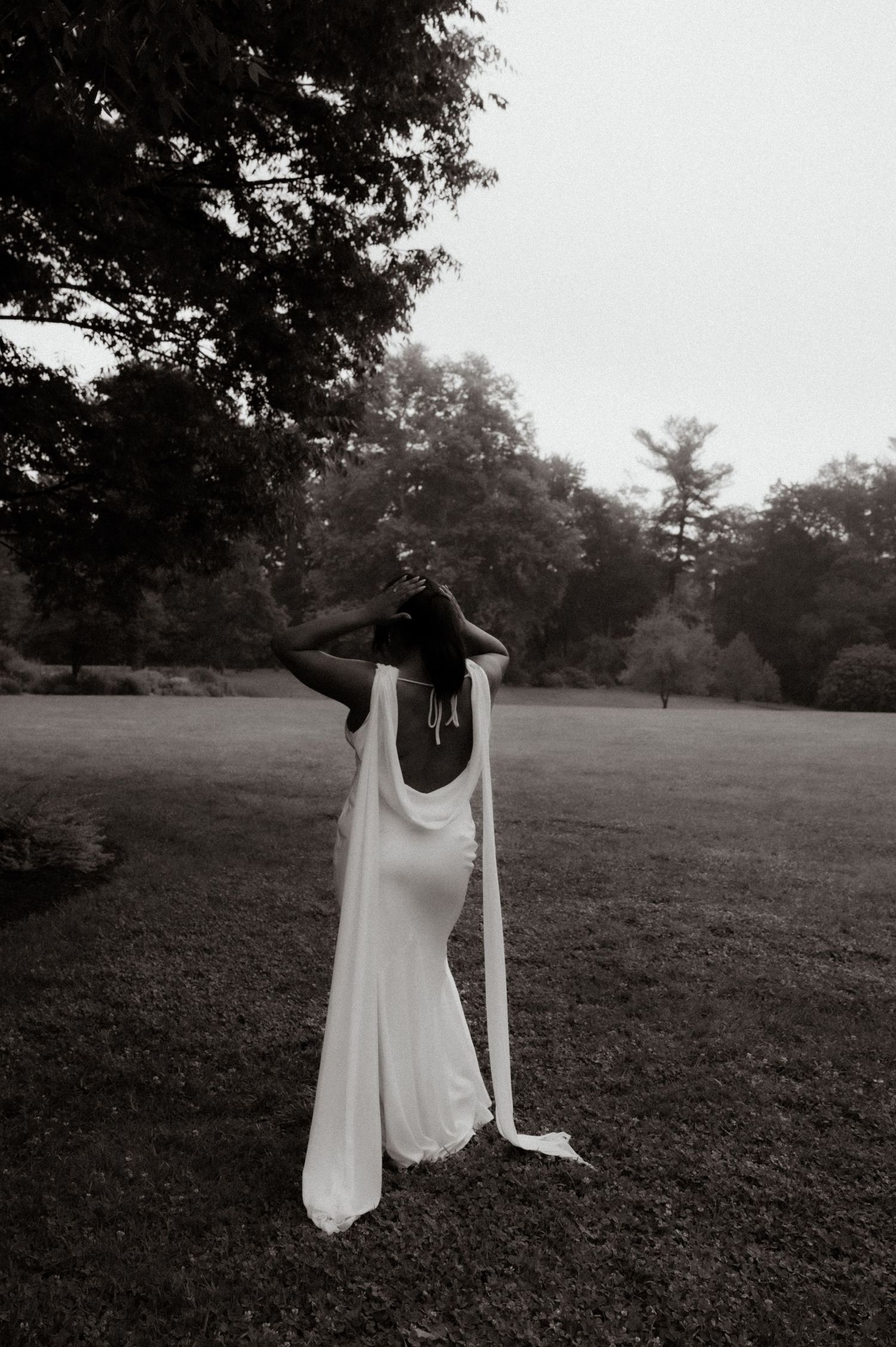 Bride in elegant white backless gown with flowing cape stands in lush green park, black and white photo.