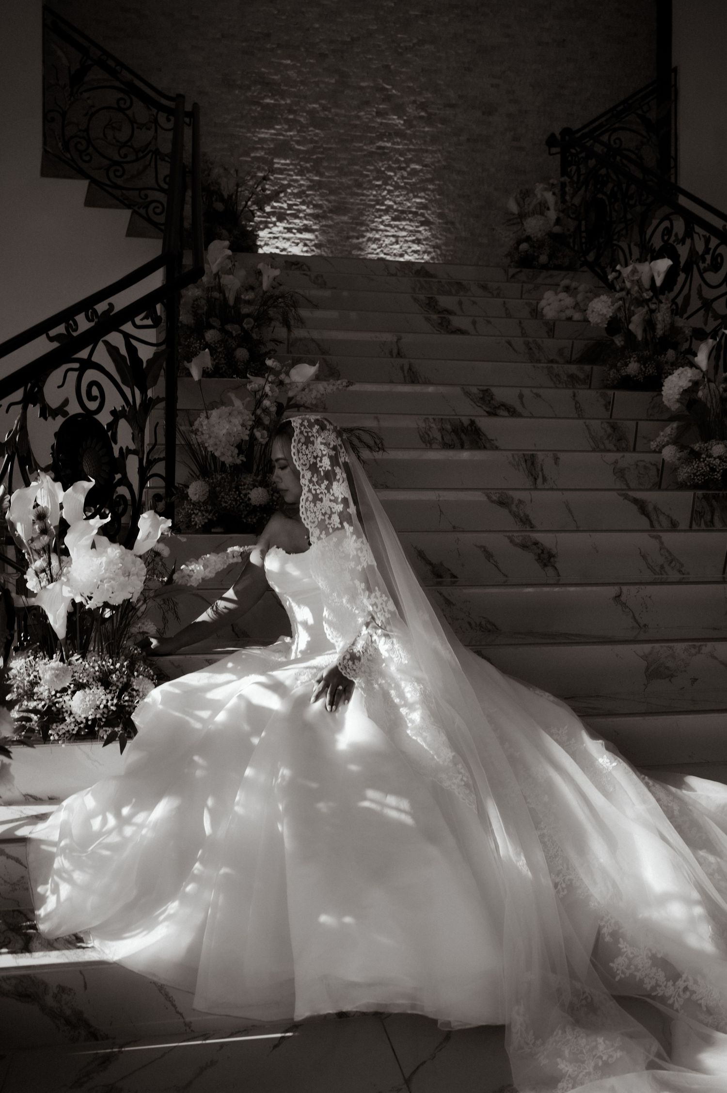 Bride in elegant white gown with long veil on marble staircase, black and white wedding photo with floral arrangements.