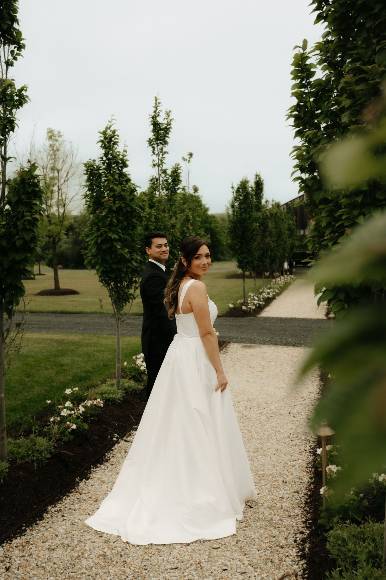 Bride in white ball gown and groom in black tuxedo pose on a garden path lined with trees and flowers.