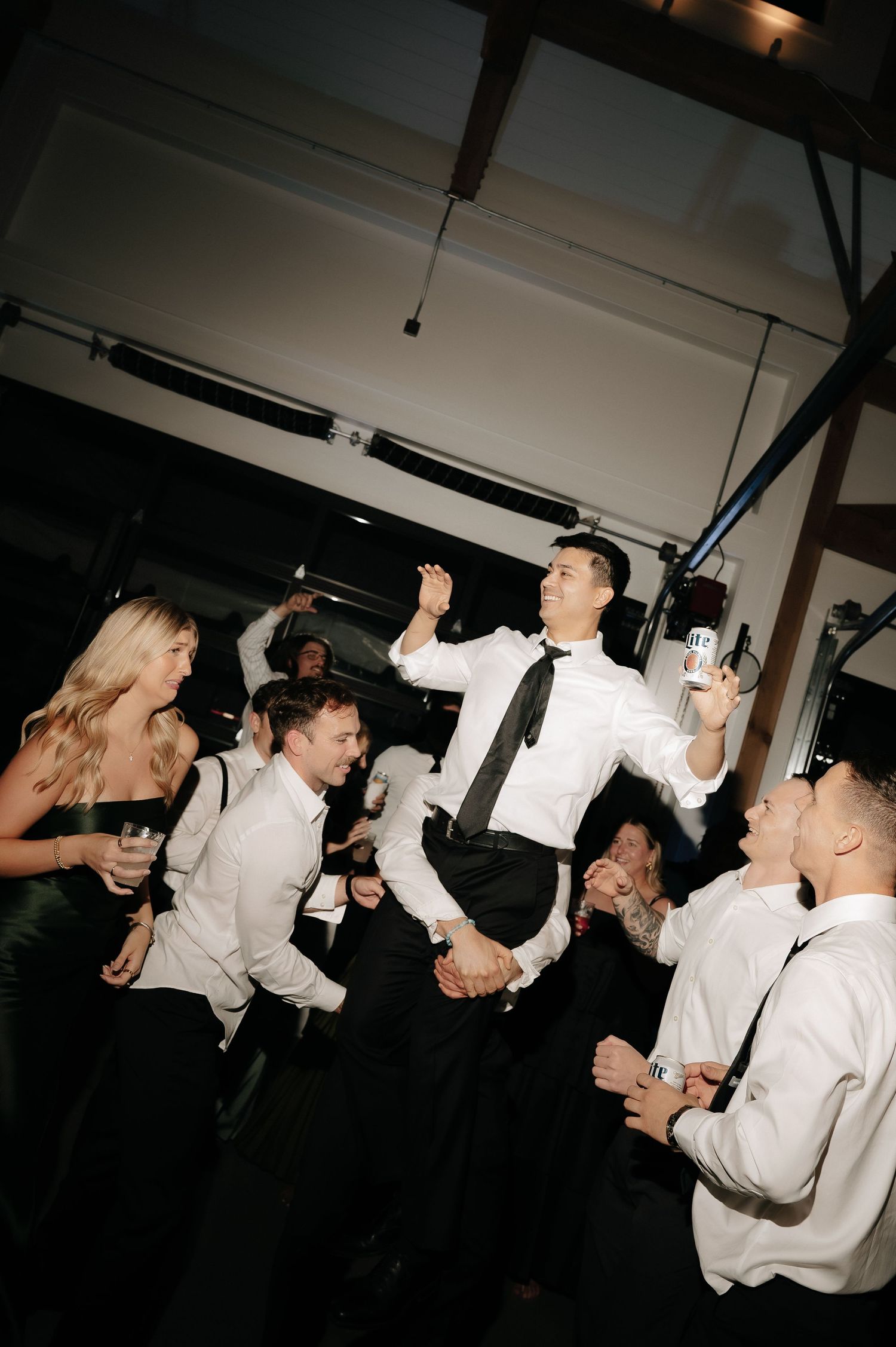 Guests dancing energetically at a wedding reception, with a man in a white shirt and black tie holding a bottle.
