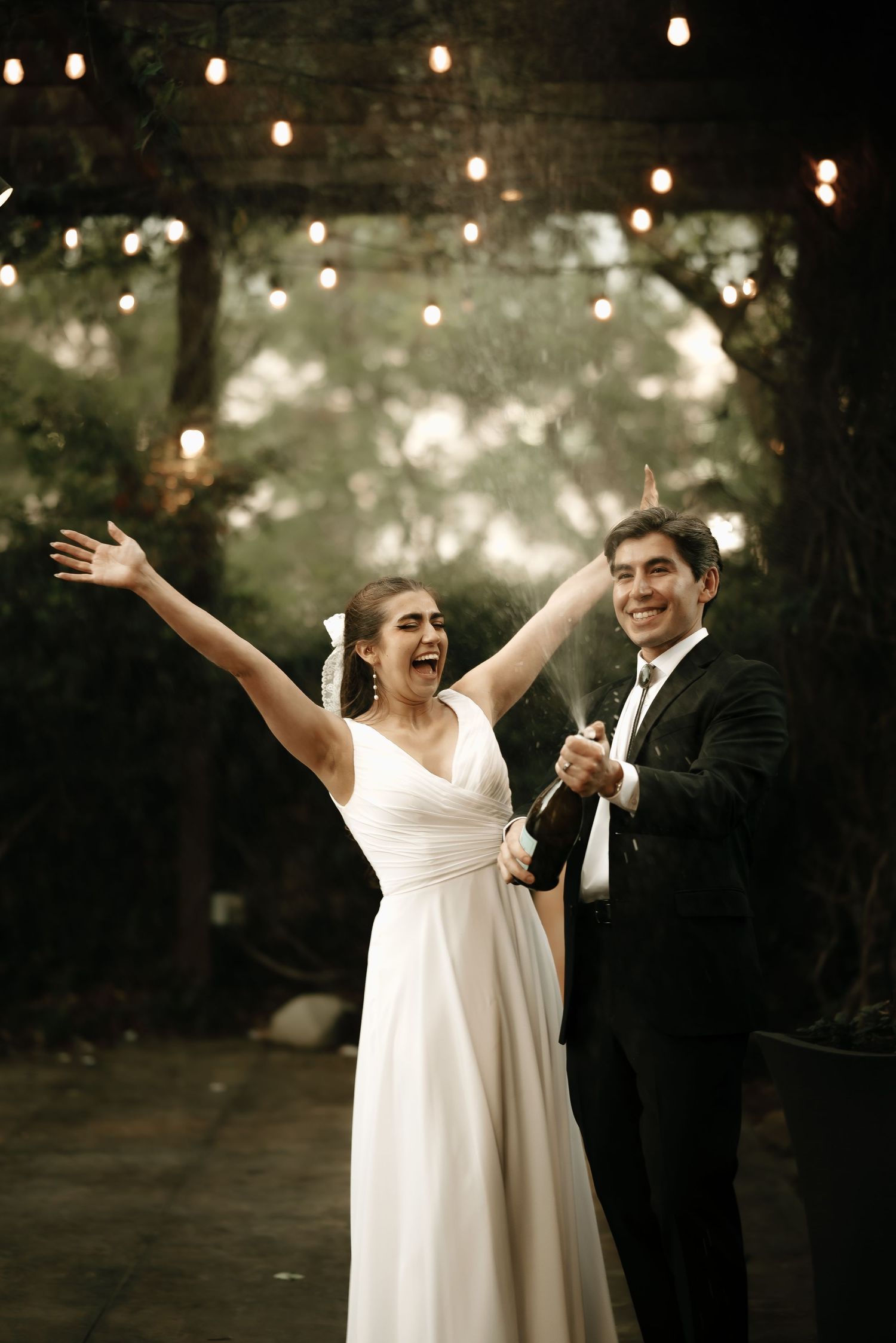 Wedding couple dancing joyfully under twinkling outdoor lights at dusk.