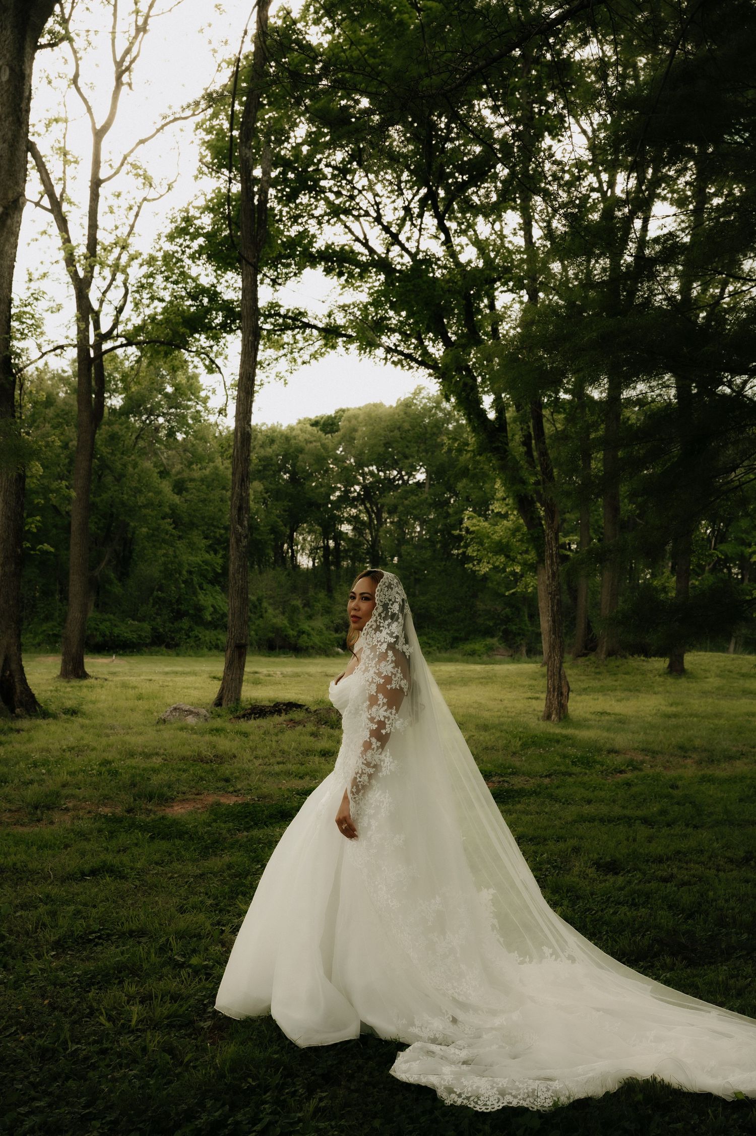 Bride in a white ball gown with lace veil stands gracefully in a lush green forest setting.