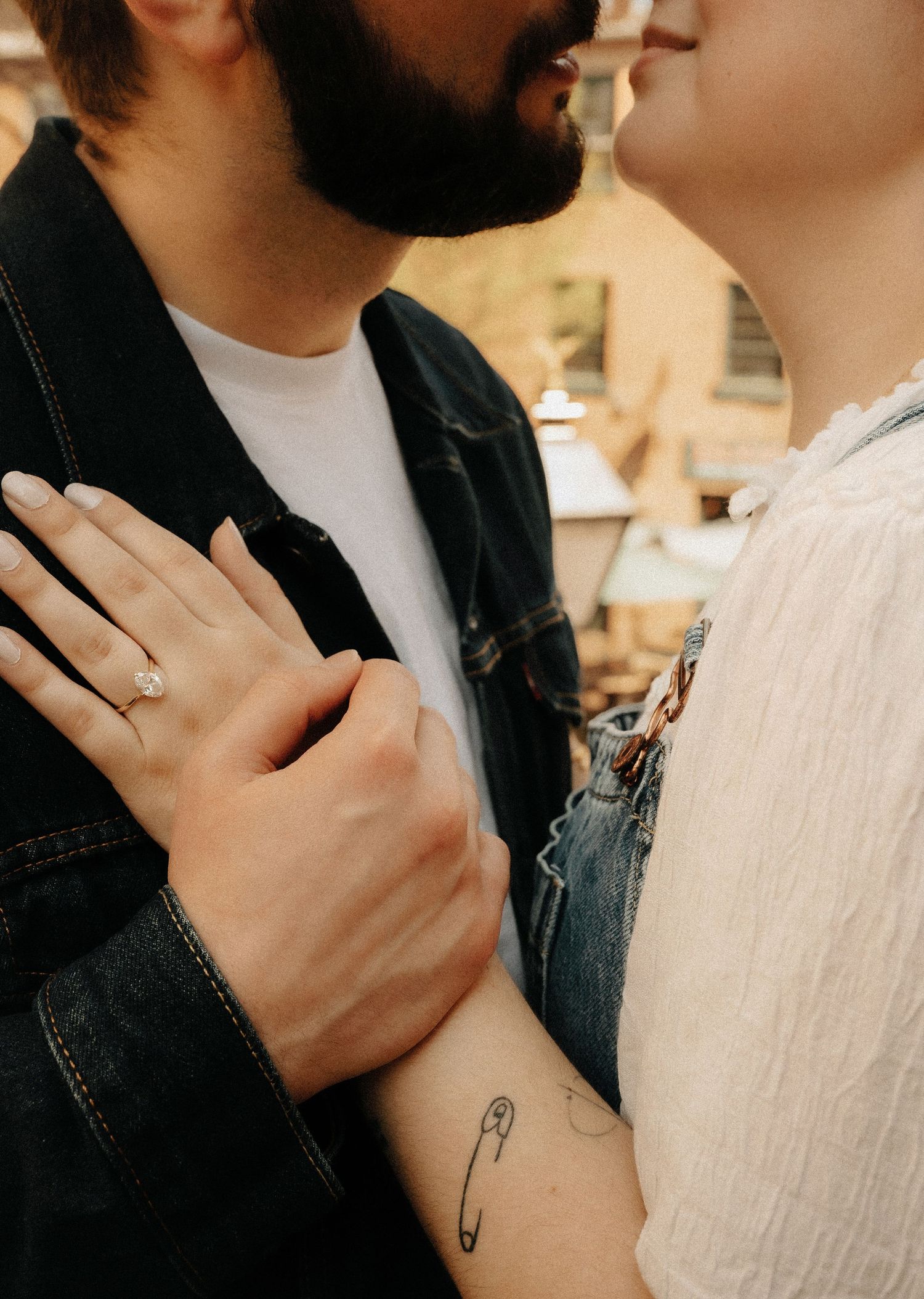 Couple nearly kissing outdoors, woman displaying diamond engagement ring and a safety pin tattoo on her wrist.