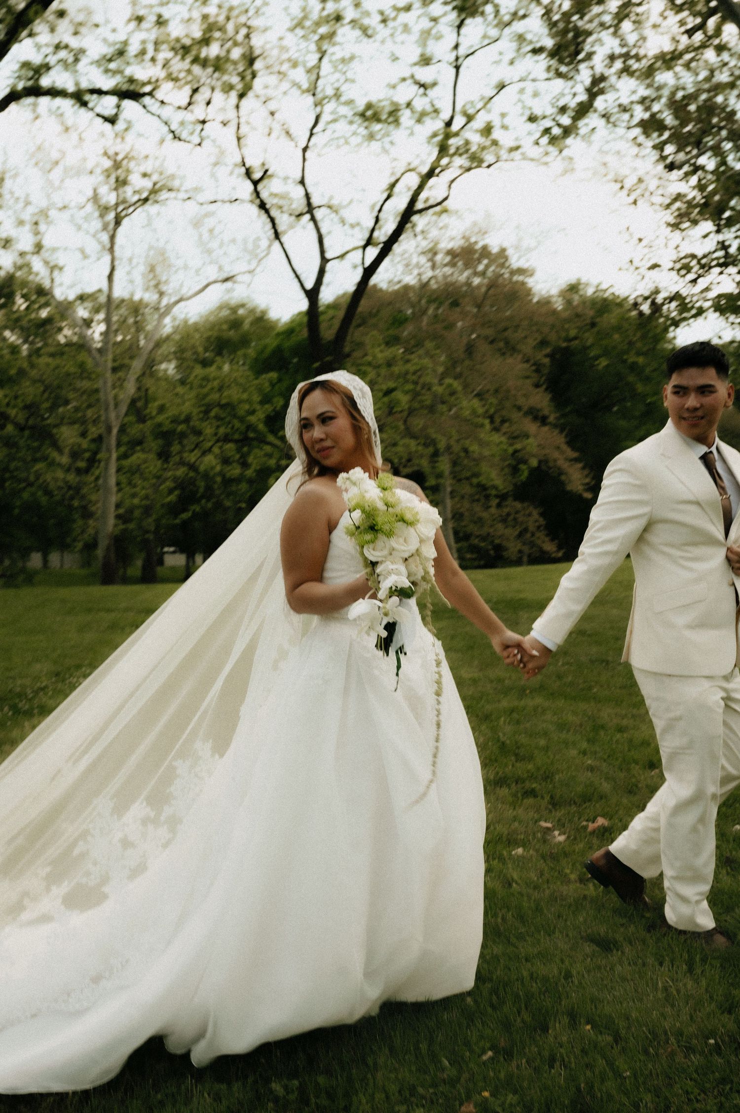 Bride in white gown and veil holds bouquet while walking hand-in-hand with groom in white suit in lush green park.