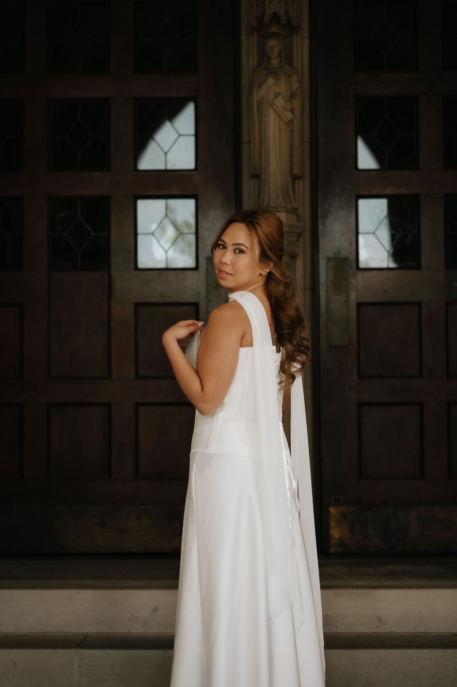 A woman in a white gown poses elegantly in front of ornate wooden doors with stained glass windows.