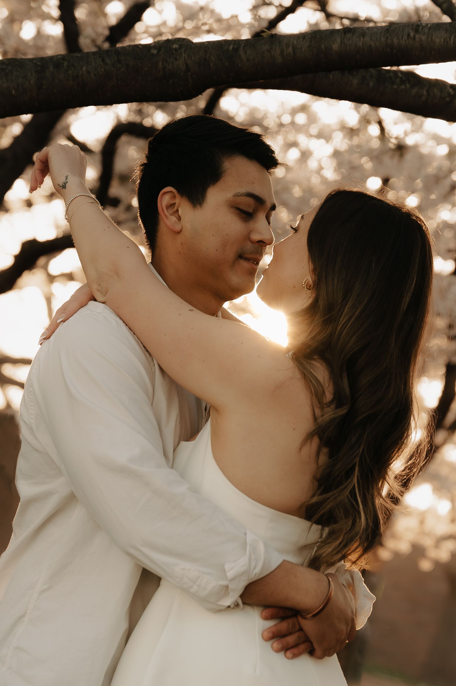 Couple embracing and almost kissing outdoors at golden hour under blooming trees, wearing white outfits.