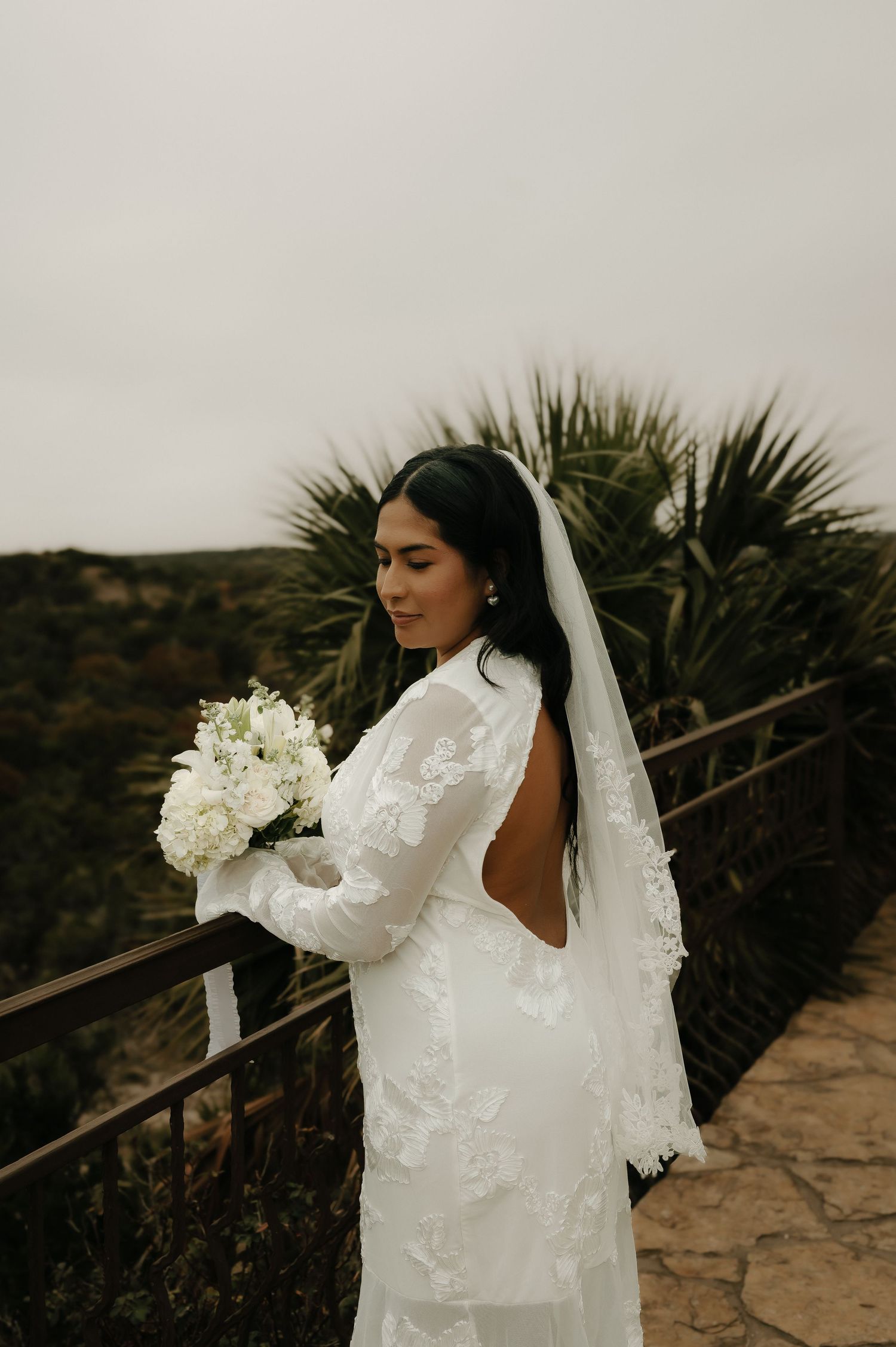 Bride in long-sleeved lace wedding dress holding white bouquet on stone terrace.