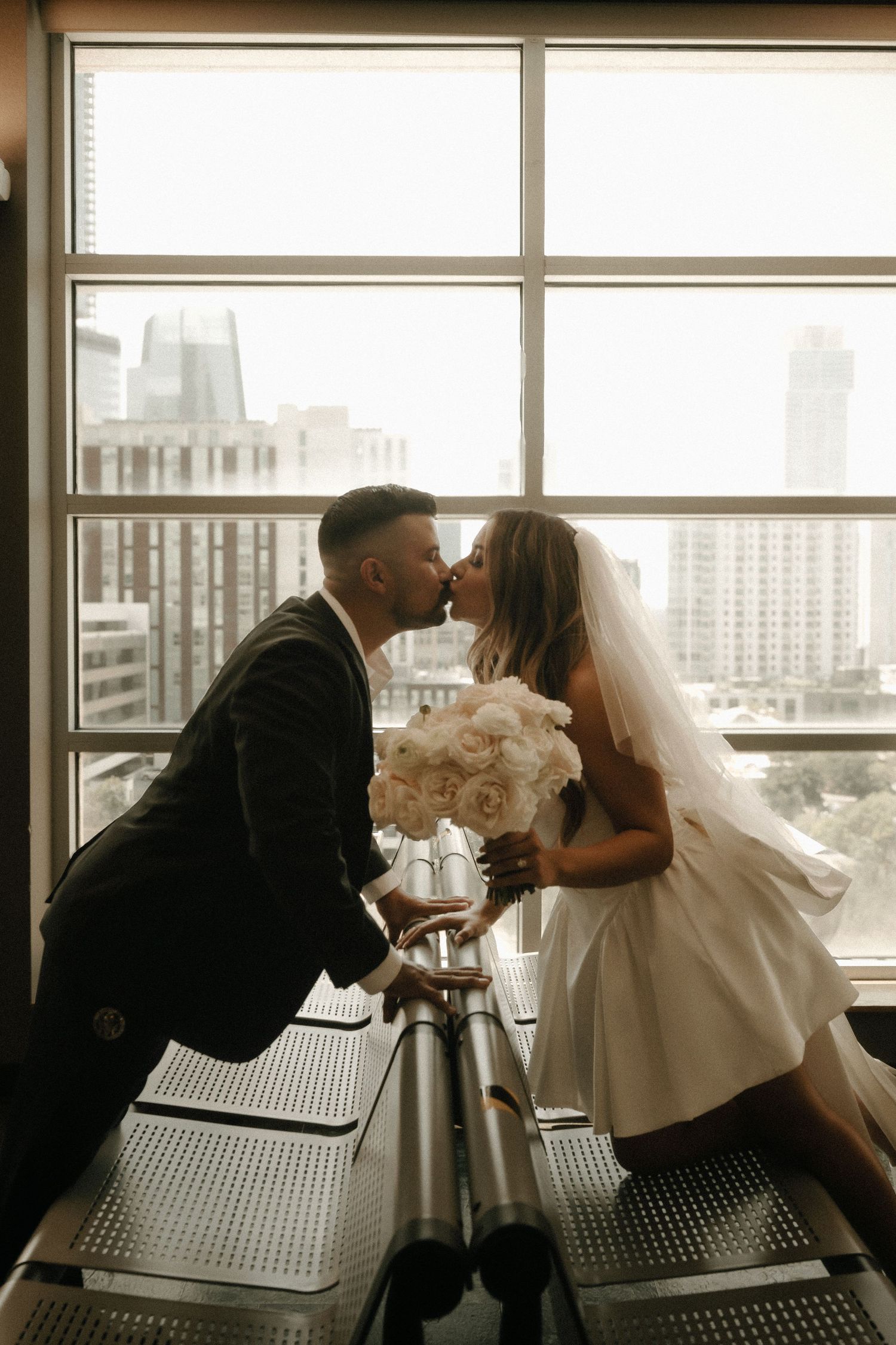 A romantic couple shares a kiss in front of large windows overlooking a city skyline.