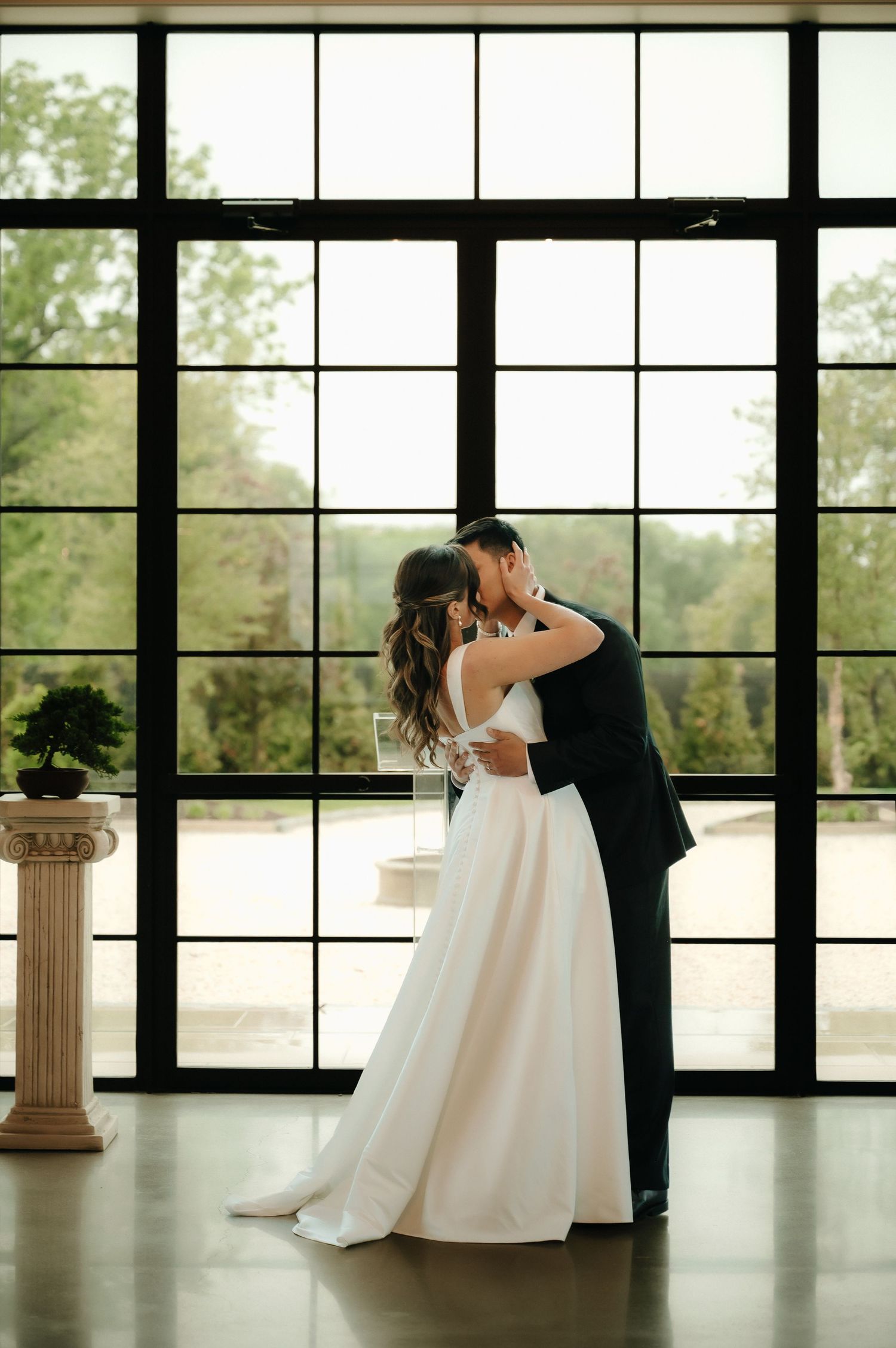 Bride and groom share a romantic first kiss in front of large black-framed windows at an elegant wedding venue.