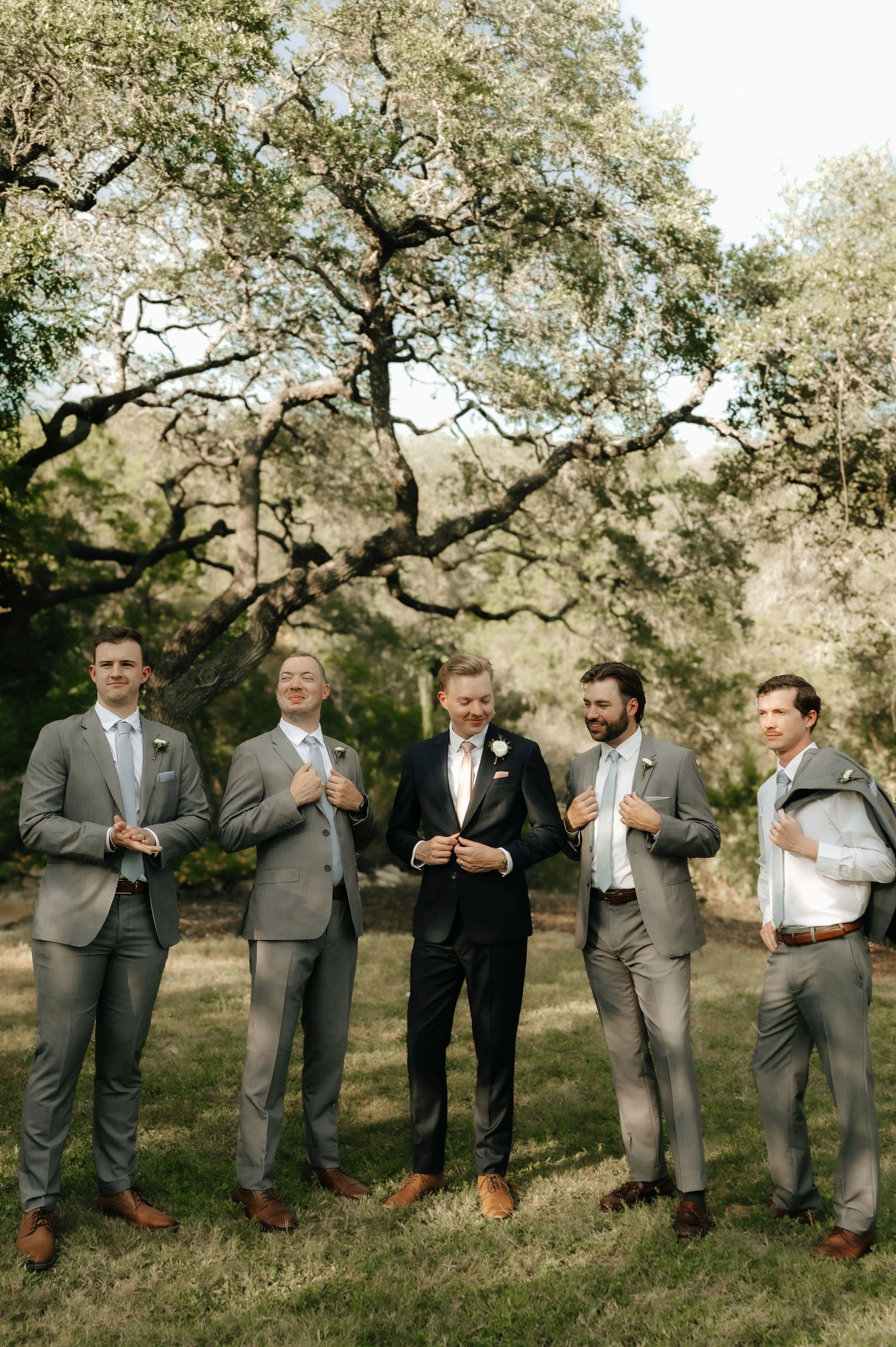 Groomsmen in grey suits standing casually on grassy hillside under oak trees.