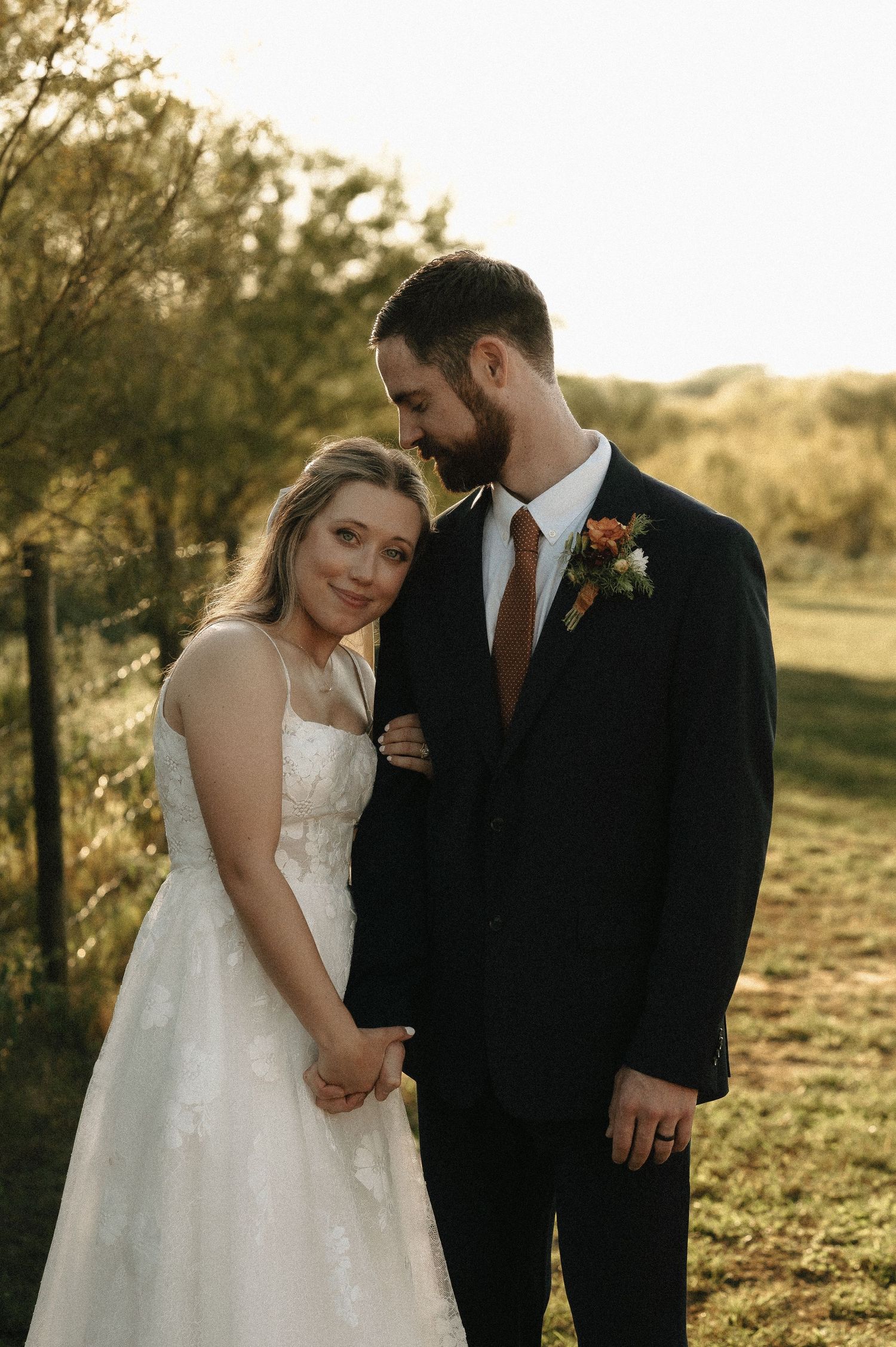 A romantic sunset wedding portrait of a couple in formal attire standing in a golden-lit field.