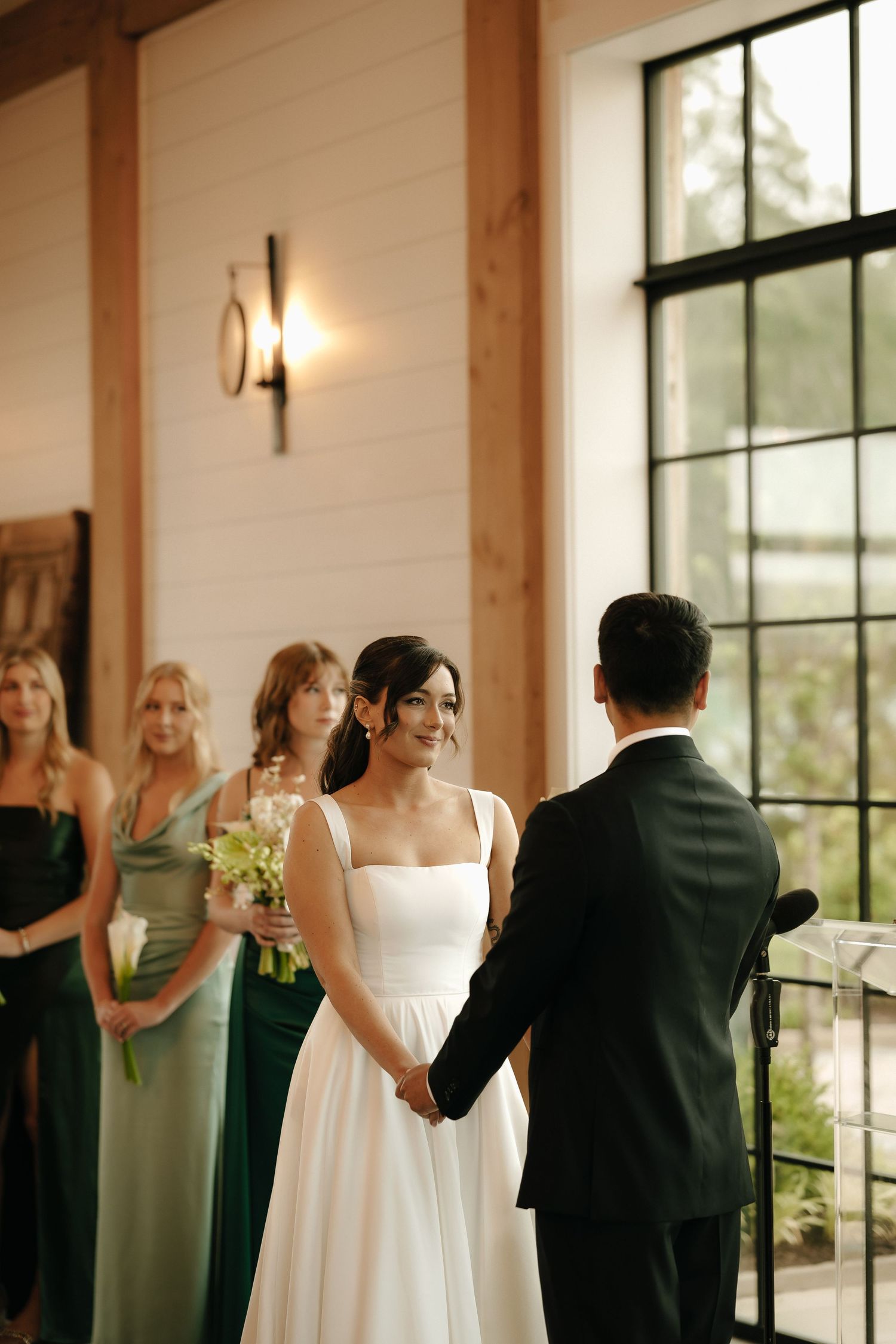 Bride in white gown holds hands with groom at indoor wedding ceremony, bridesmaids in green dresses stand behind her.