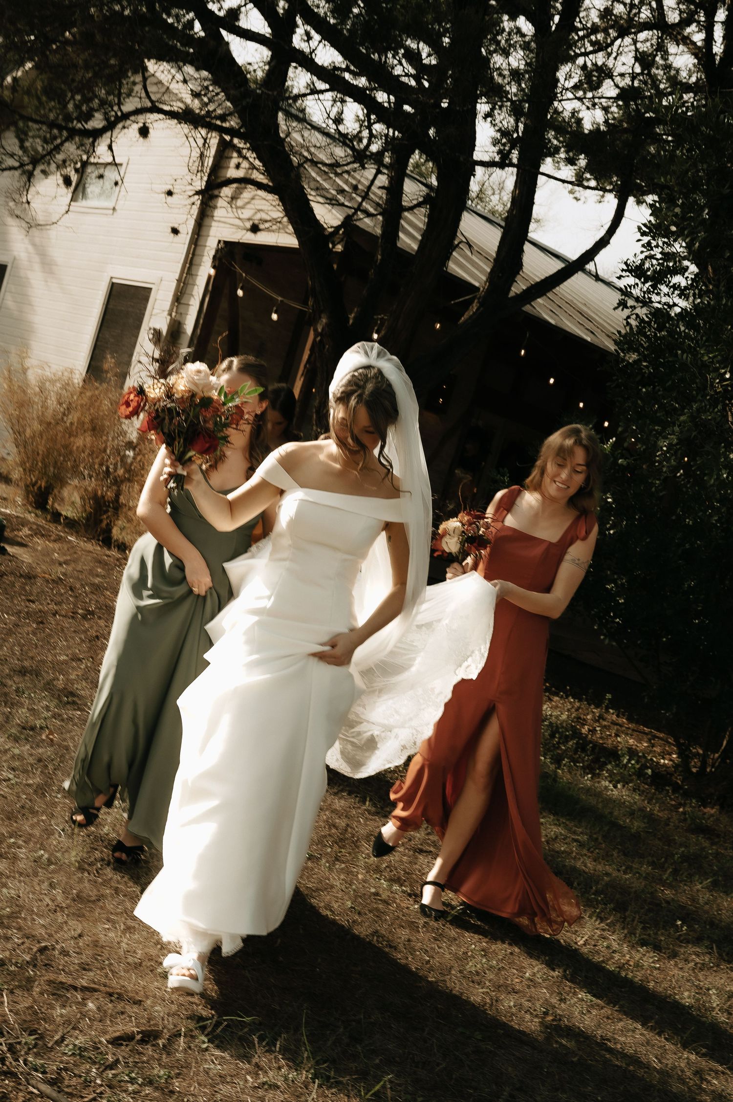 Bride in white dress walks outdoors with bridesmaids in earth-toned dresses.