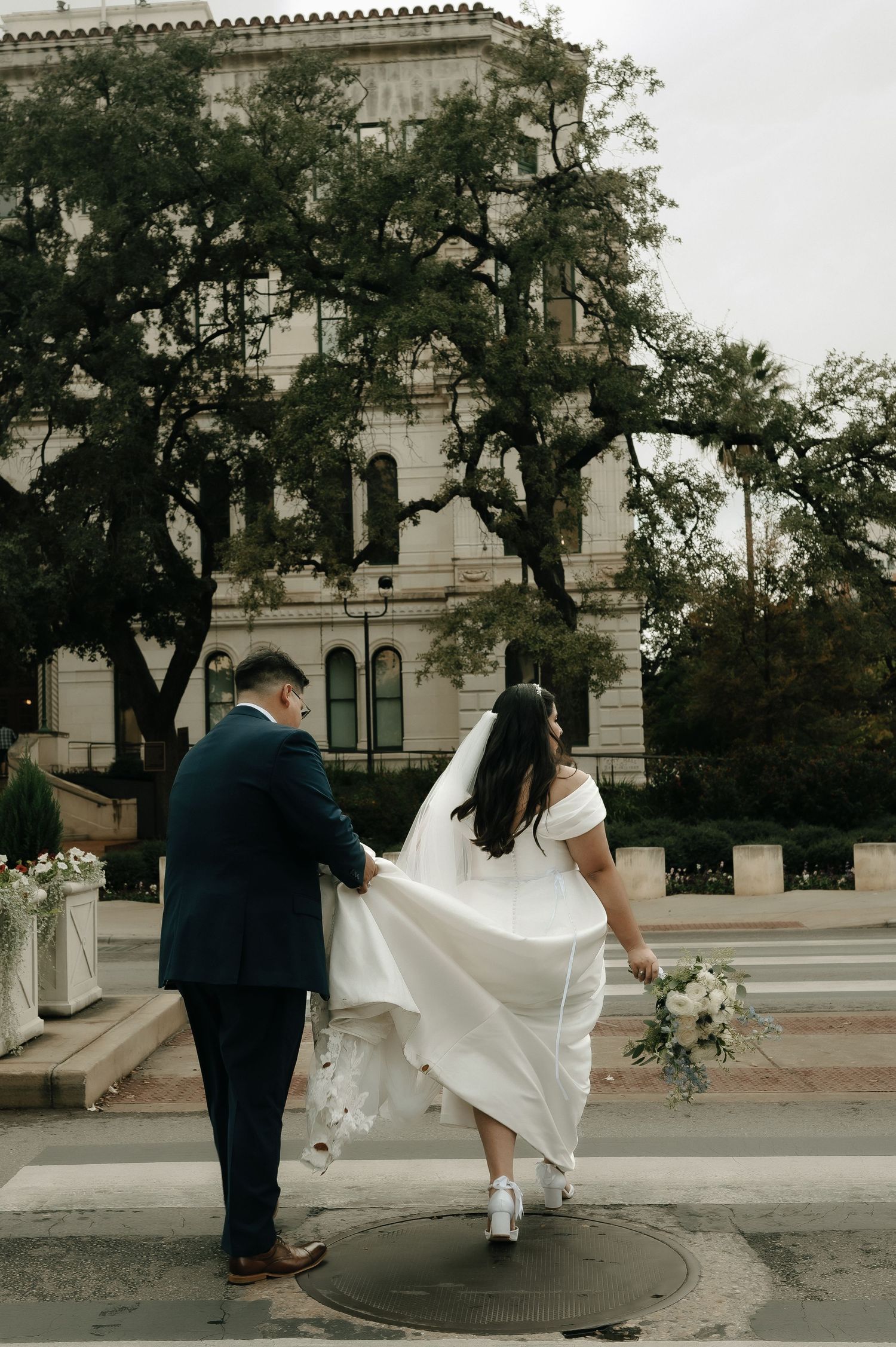 Wedding couple walks together outside historic building with flowing dress train.