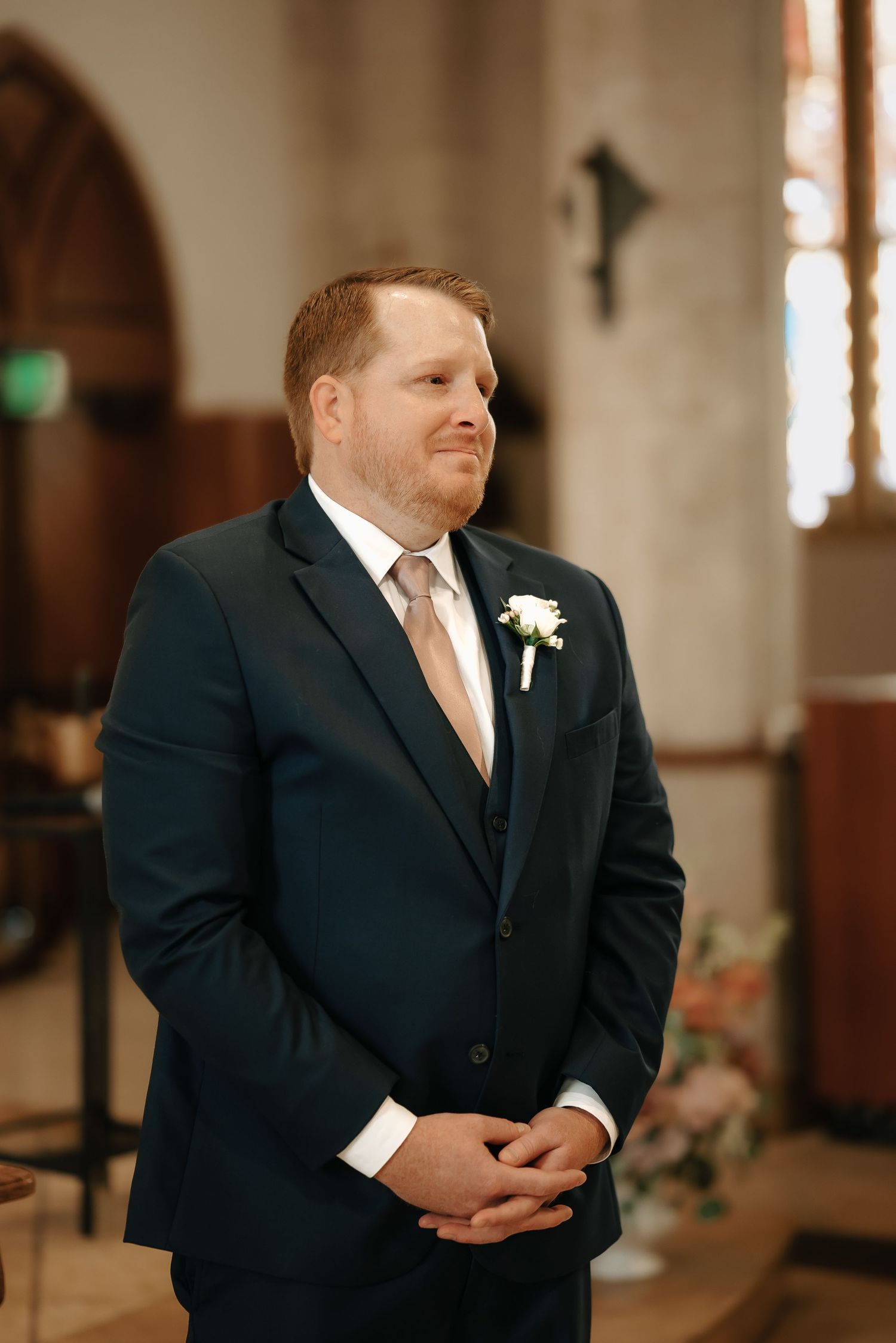 Groom in a navy suit and peach tie smiling during wedding ceremony in church.