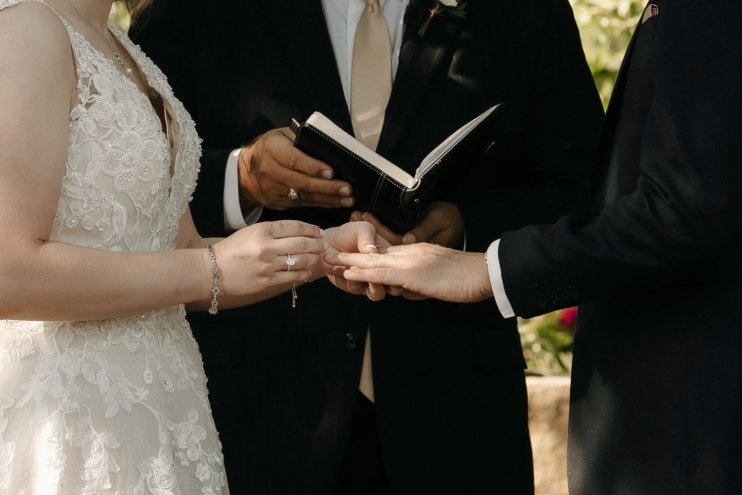 A couple exchanges vows during an outdoor wedding ceremony under green foliage.