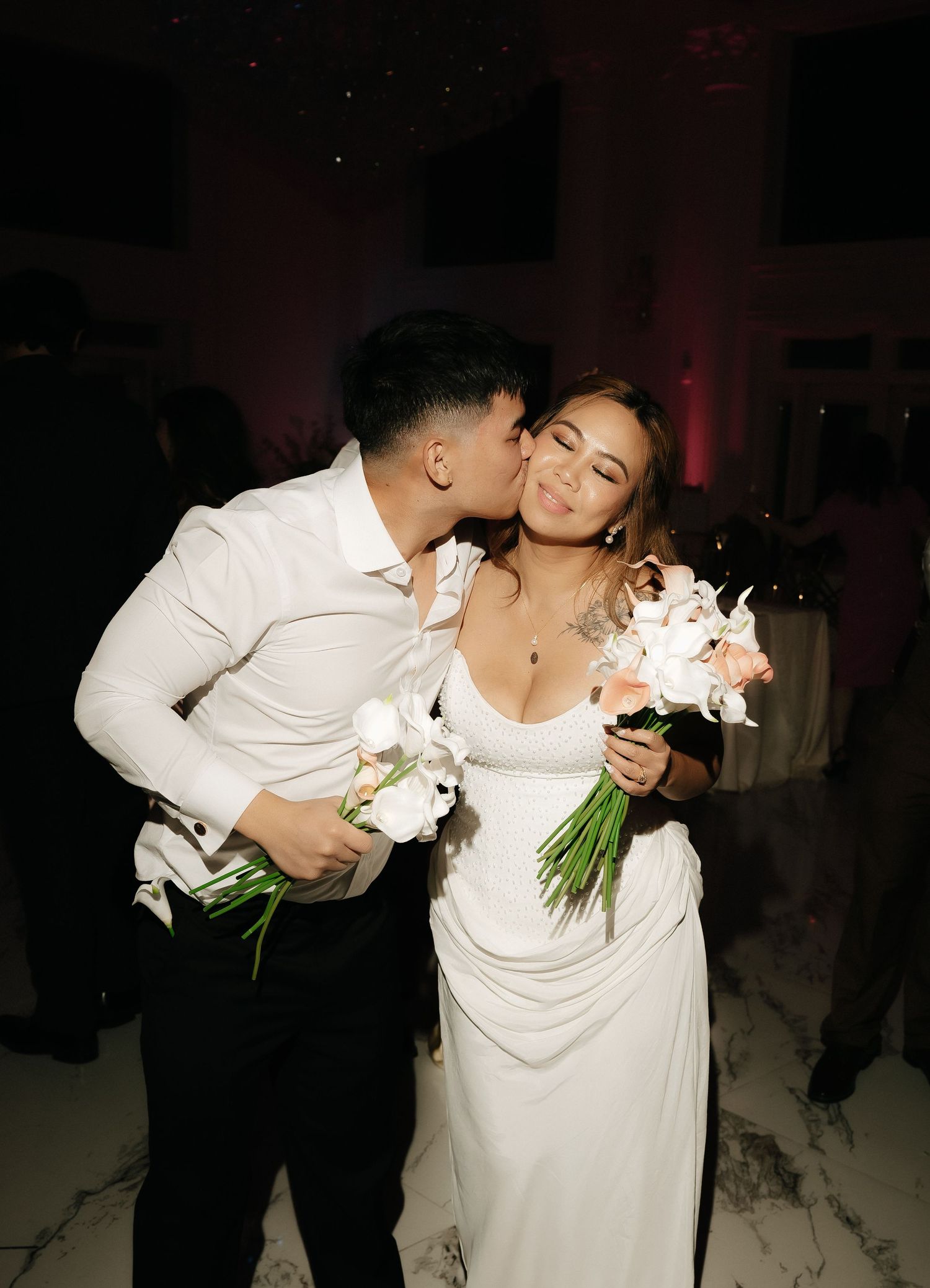 Groom kissing bride on cheek at wedding reception, bride smiling holding white floral bouquet in elegant white gown.