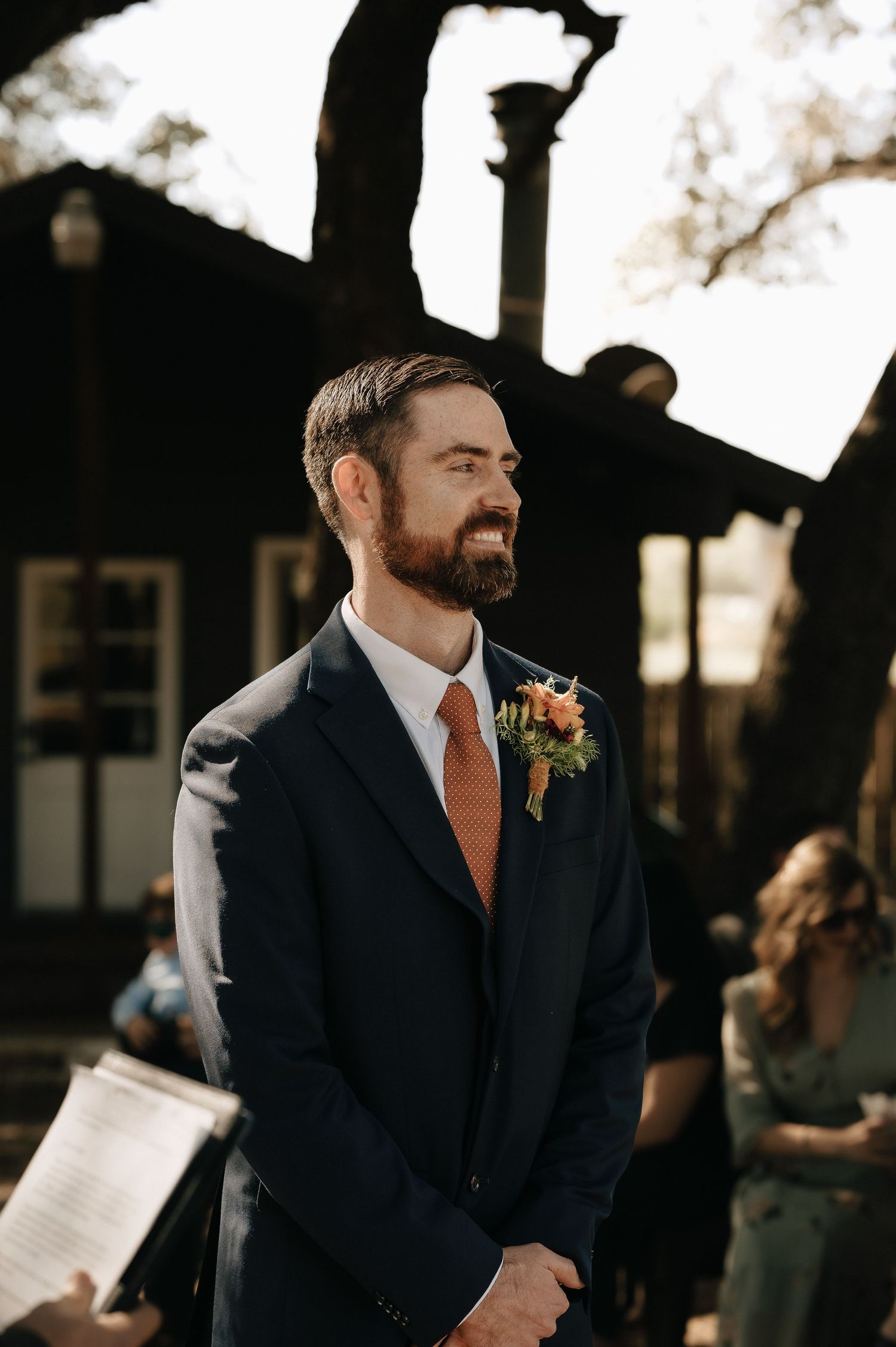 Groom in navy suit smiles during outdoor wedding ceremony.