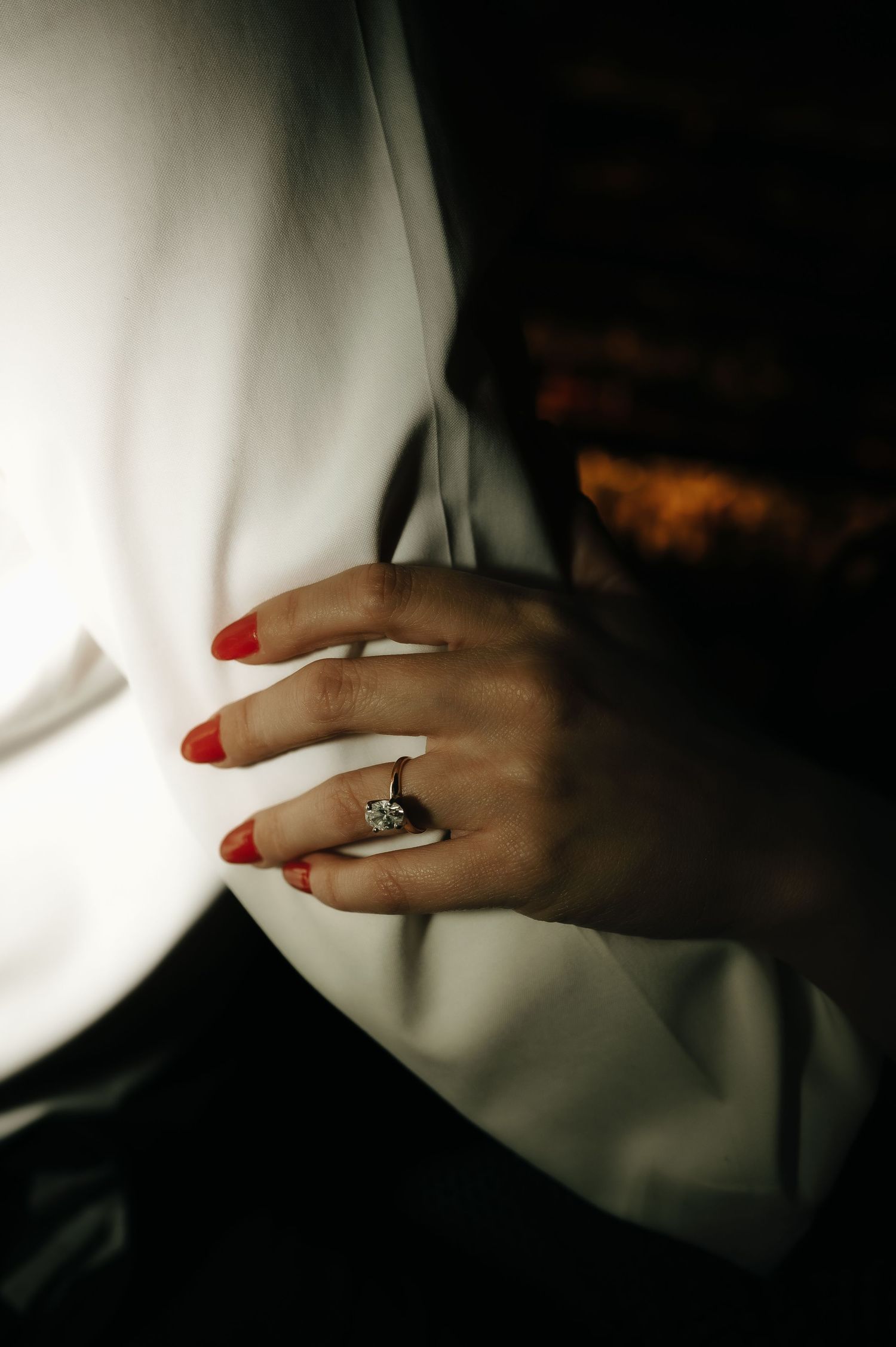 Close-up of hands with red nail polish and an engagement ring against white fabric.