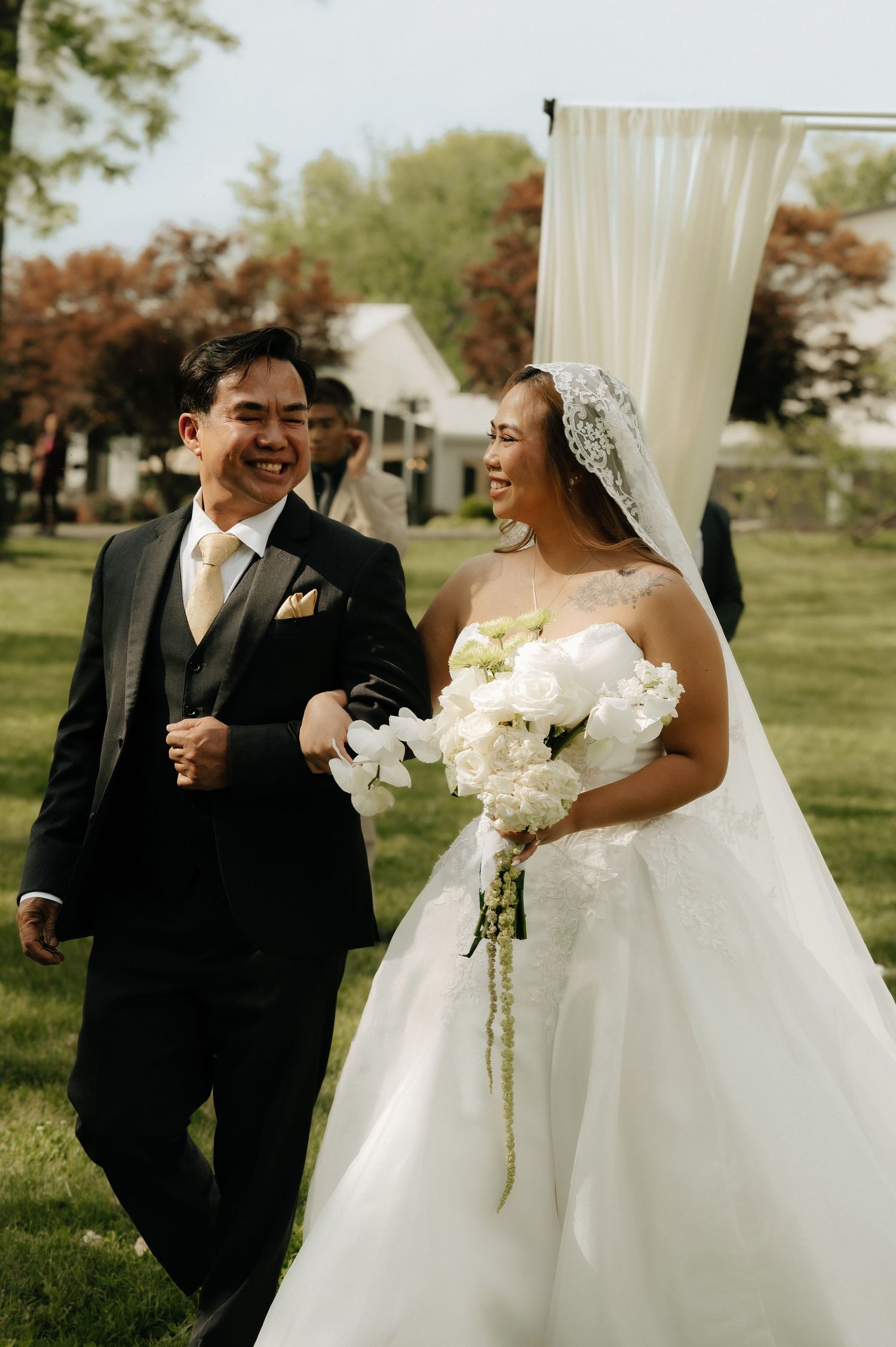 Smiling bride and groom walk together after outdoor wedding ceremony, bride holding white bouquet with lace veil.