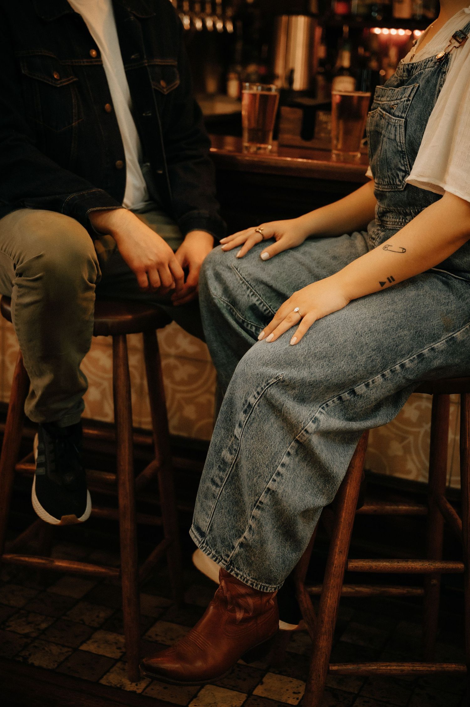 Couple sitting on bar stools holding hands, woman wearing denim overalls with engagement ring, cozy bar setting.