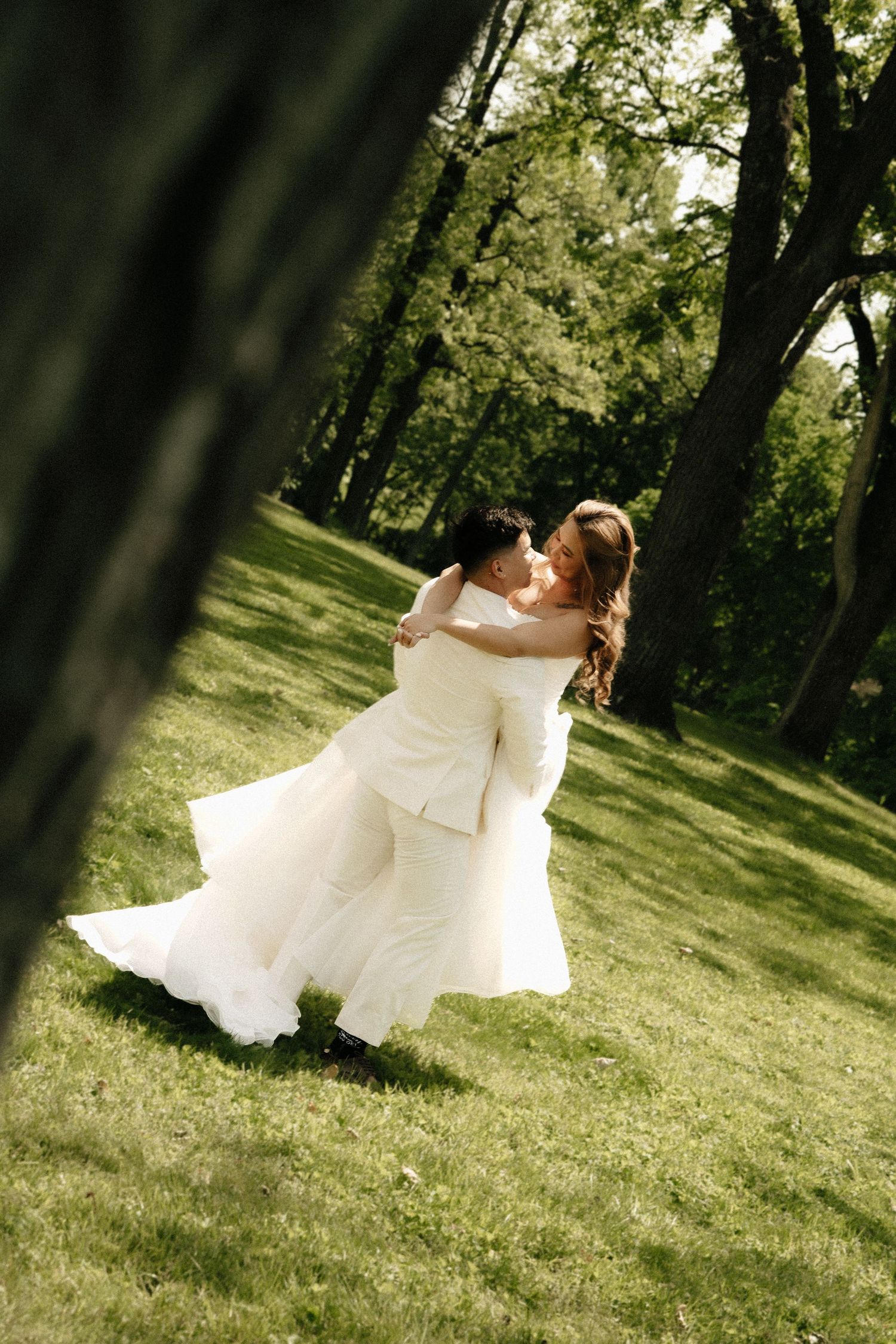 Bride in white gown lifted by groom in sunny green park, sharing romantic moment on wedding day.