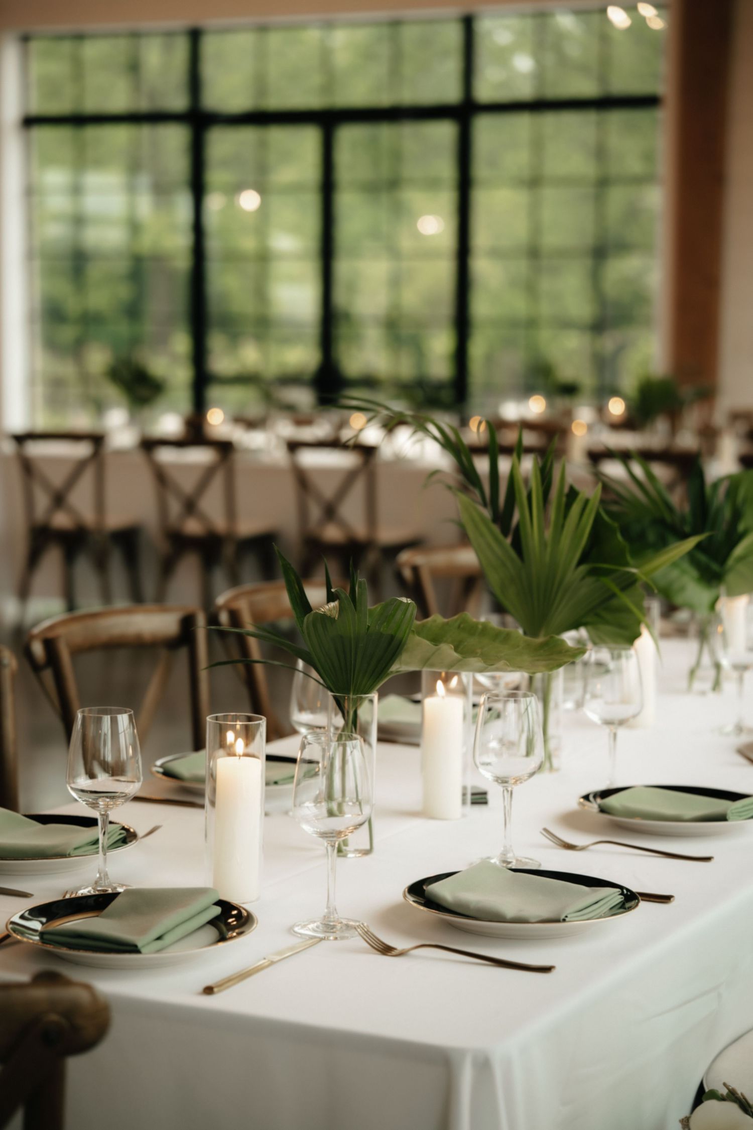 Elegant wedding reception table with white linens, candles, sage green napkins, and tropical leaf centerpieces in a bright venue.