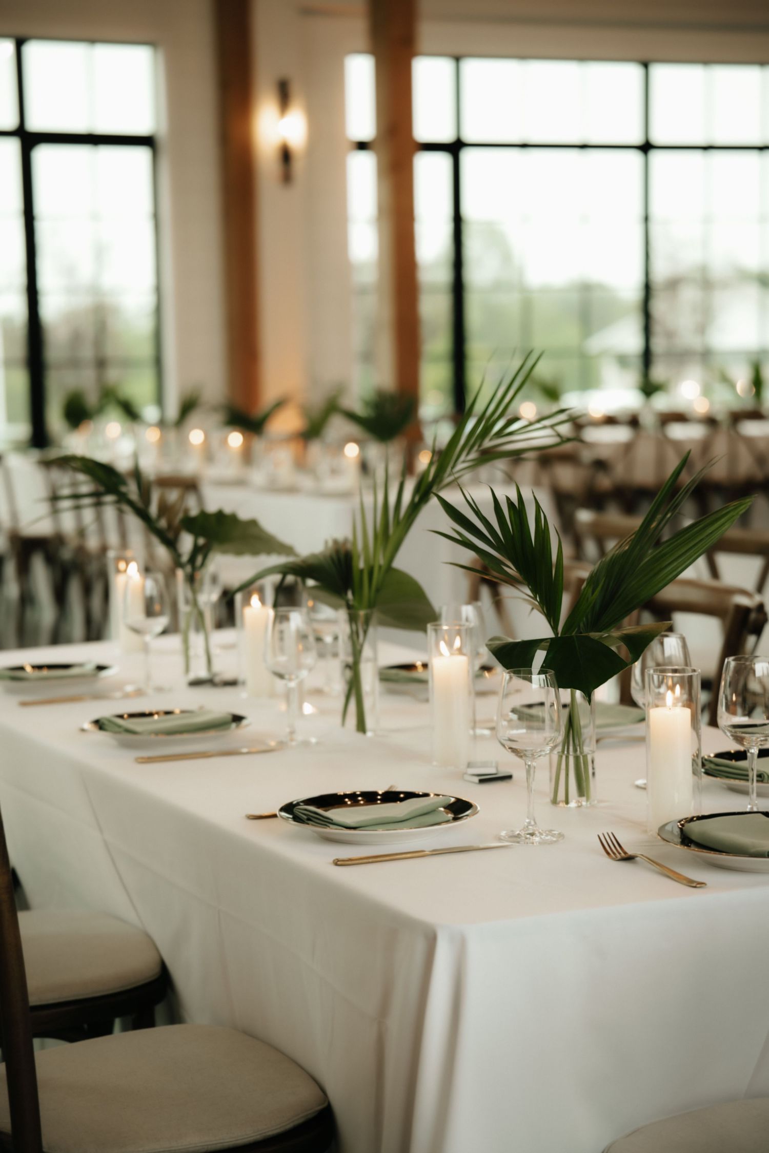Elegant wedding reception table with white linen, tropical palm leaves, candles, and green charger plates in a bright venue.