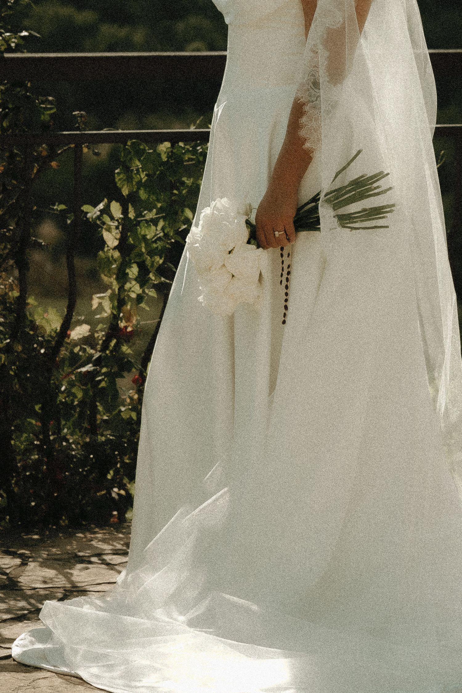 A close-up of a bride's white wedding dress and bouquet against a garden backdrop.