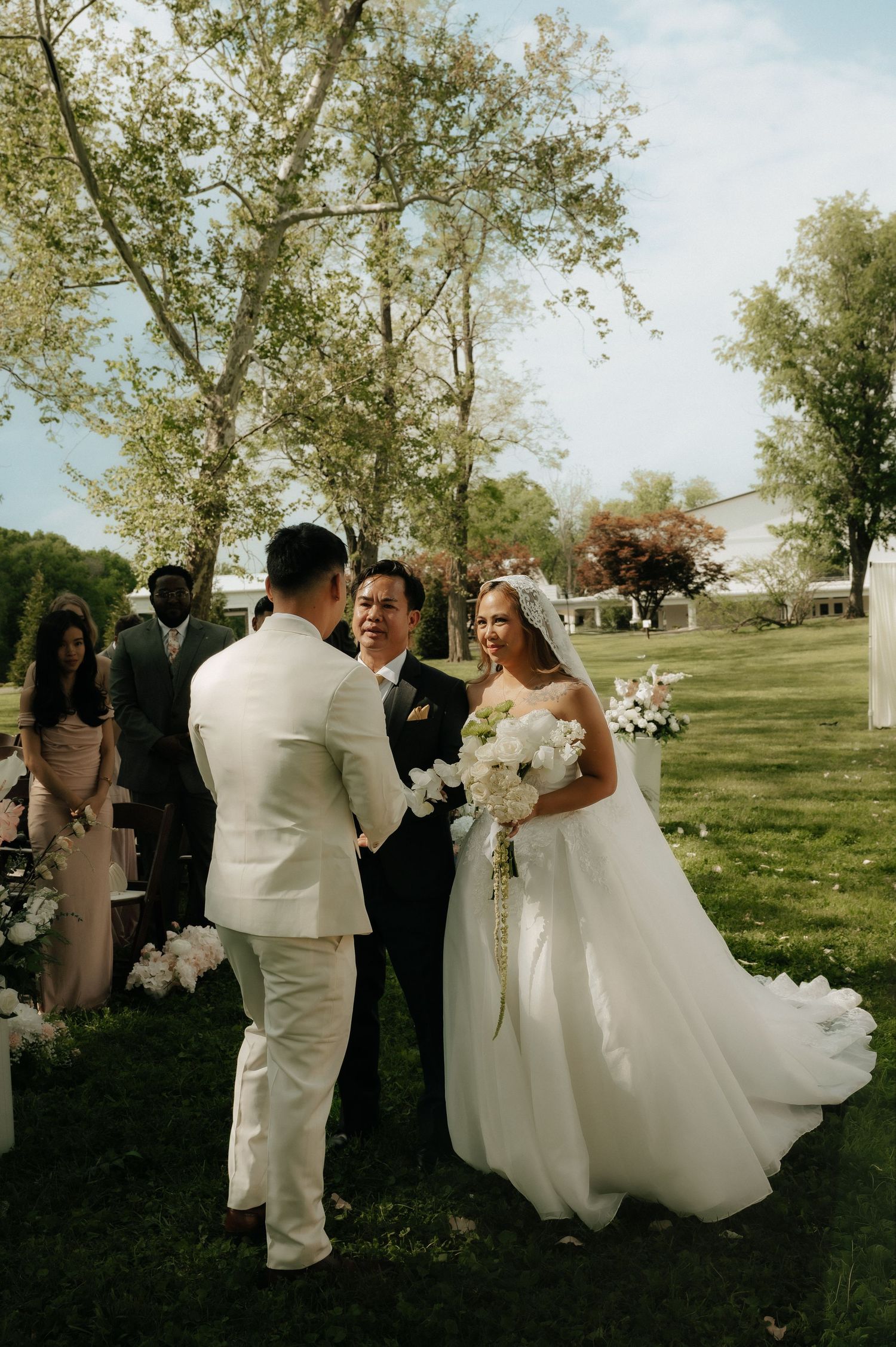 Bride in white gown holds bouquet facing groom in white suit during outdoor wedding ceremony on green lawn.